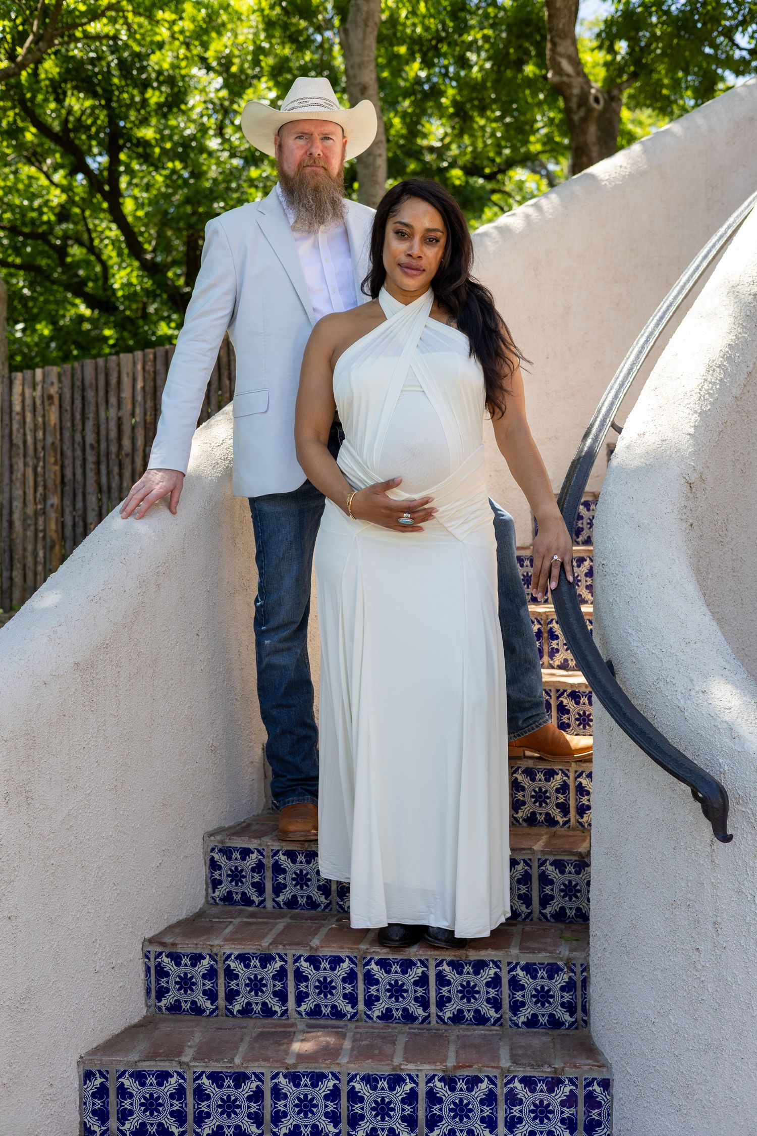 Pregnant woman in white halter dress with bearded man in cowboy hat on decorative tiled steps outdoors.