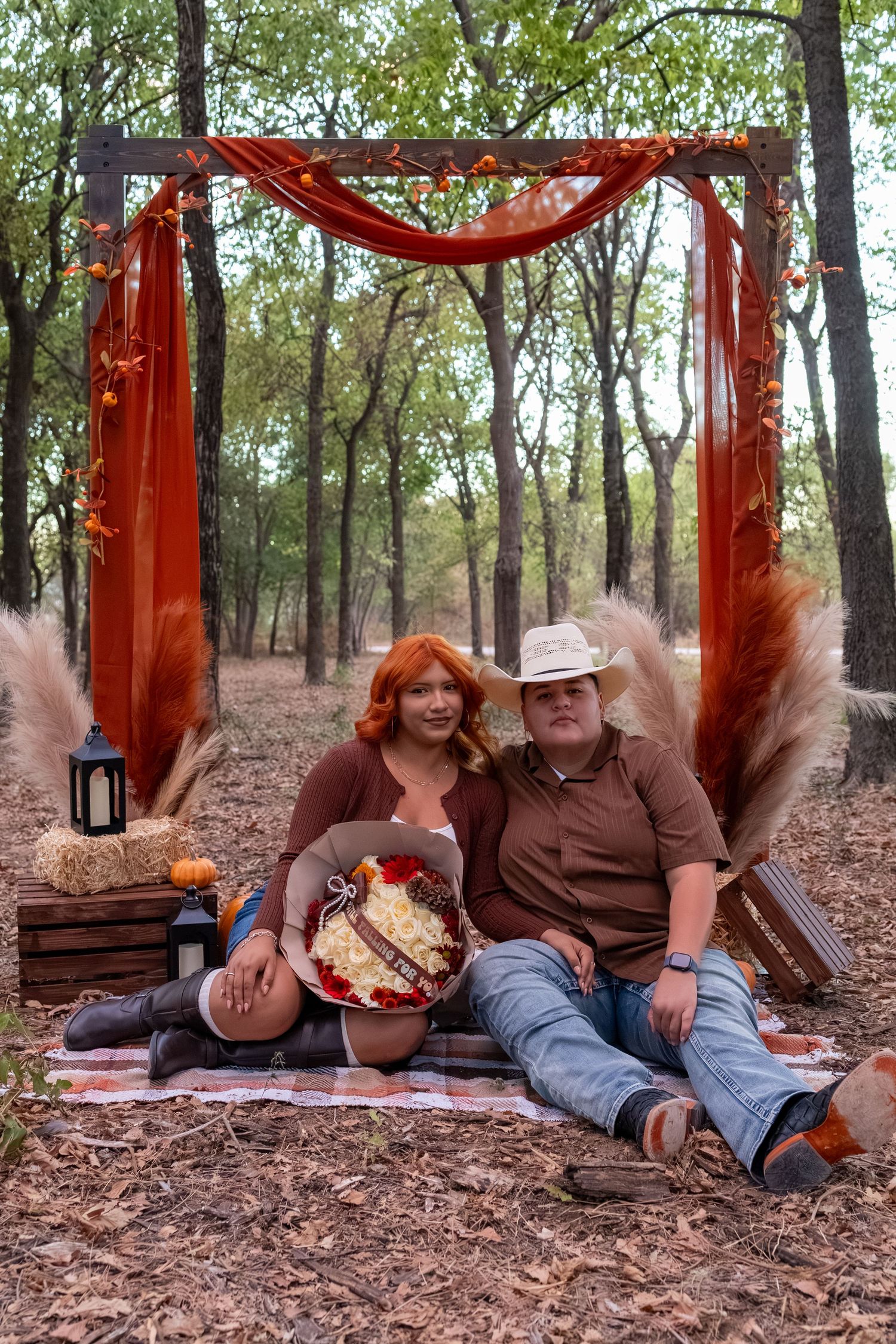 Couple sitting under rustic wooden arch with fall decor in forest, woman holding bouquet, man in cowboy hat.