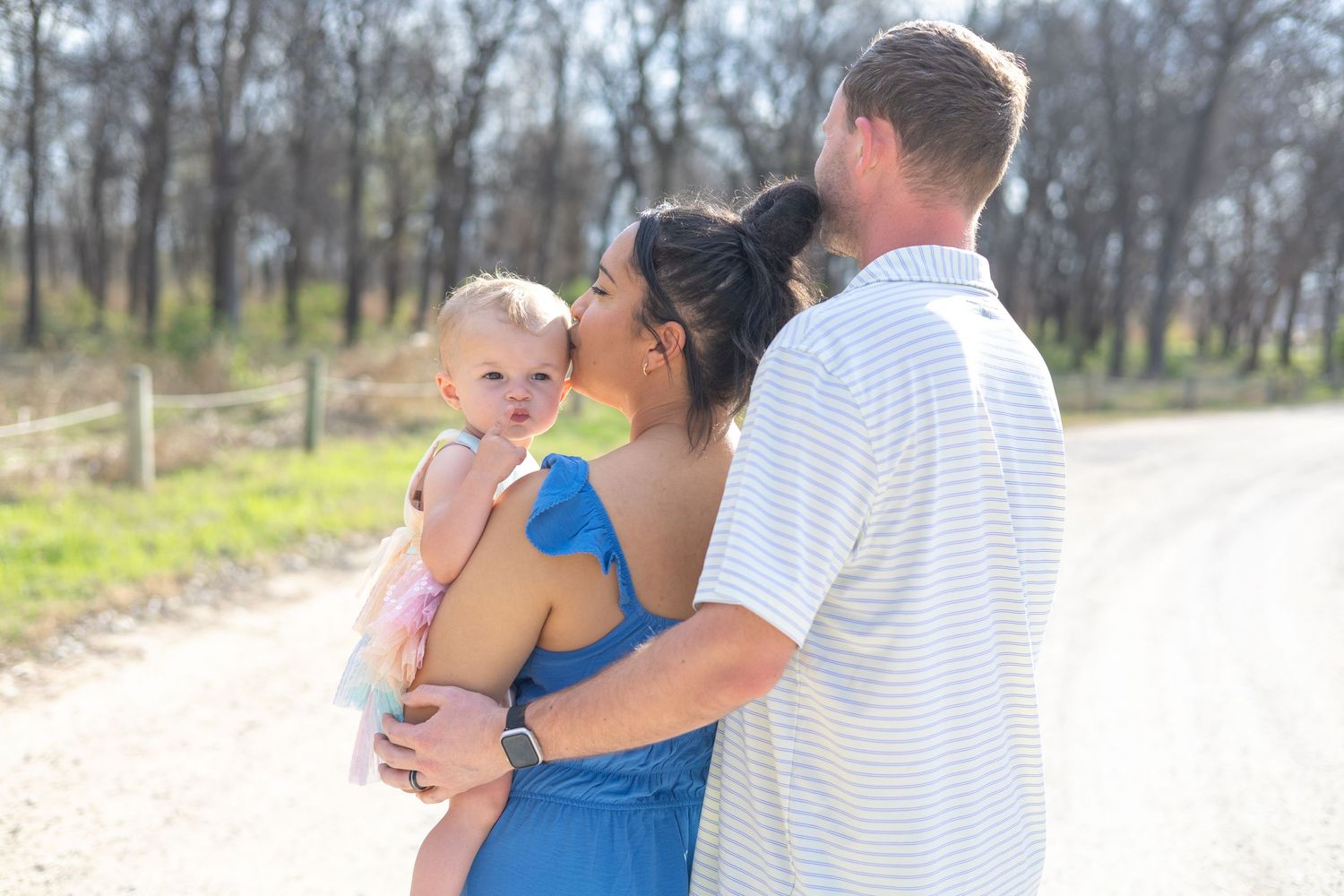 A family stands together on a sunny dirt path surrounded by trees on a warm spring day.