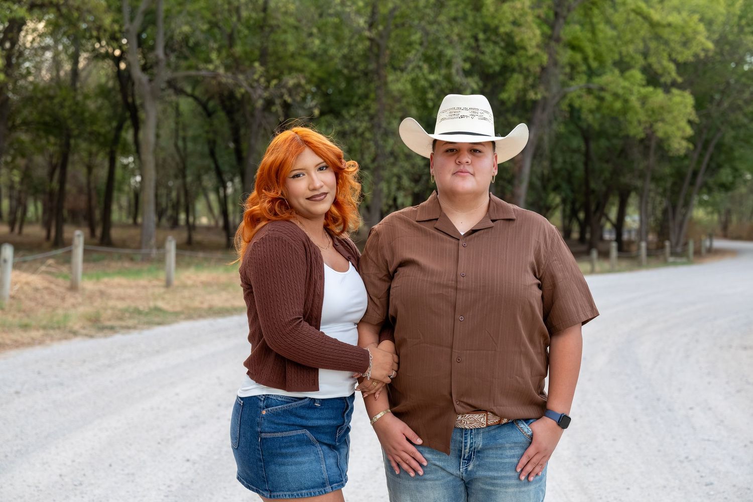 A couple poses on a rural road, woman with red hair in denim skirt, man in cowboy hat and brown shirt.