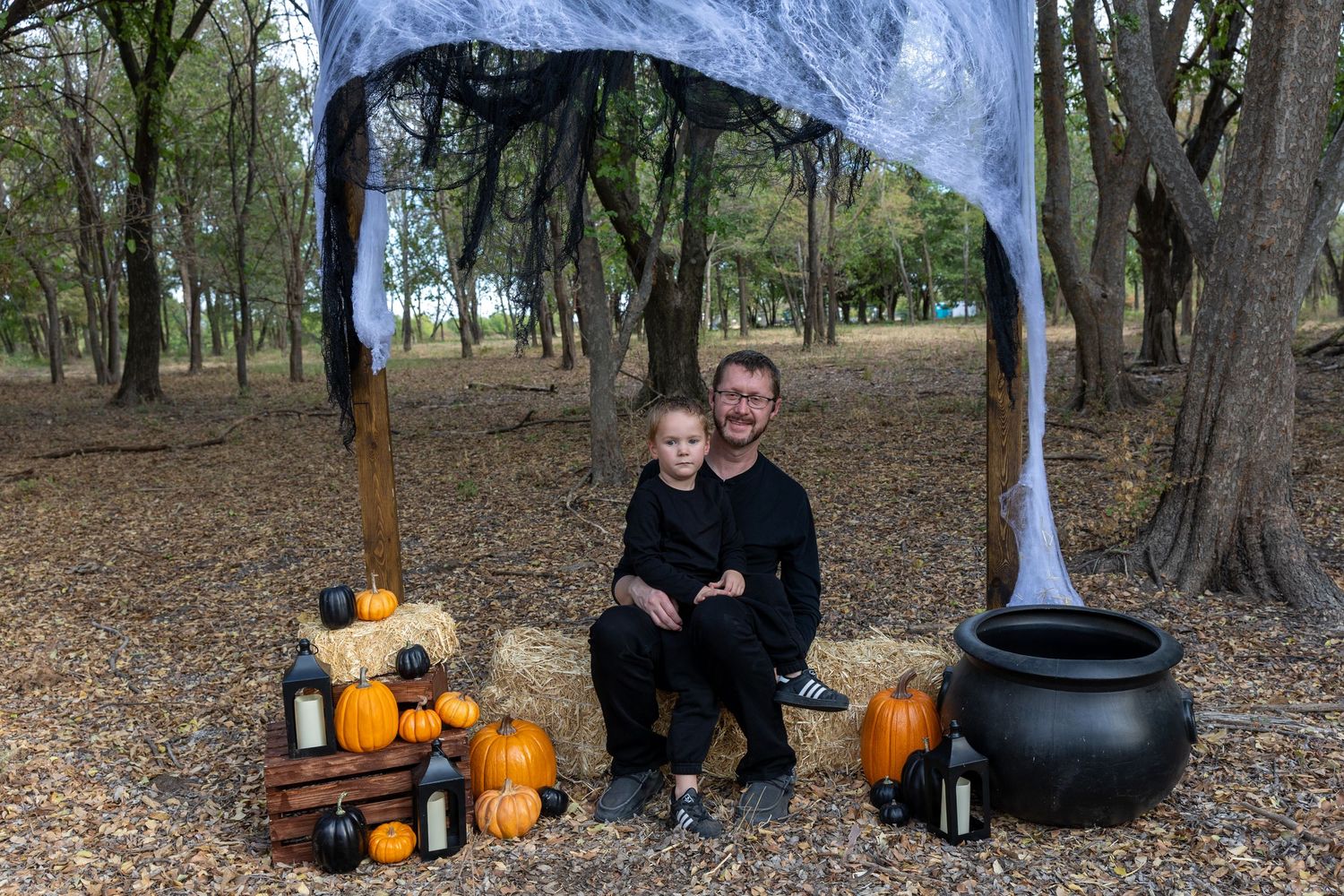 Man and child in black outfits pose at Halloween display with pumpkins, hay bales, cauldron, and spooky arch in wooded setting.