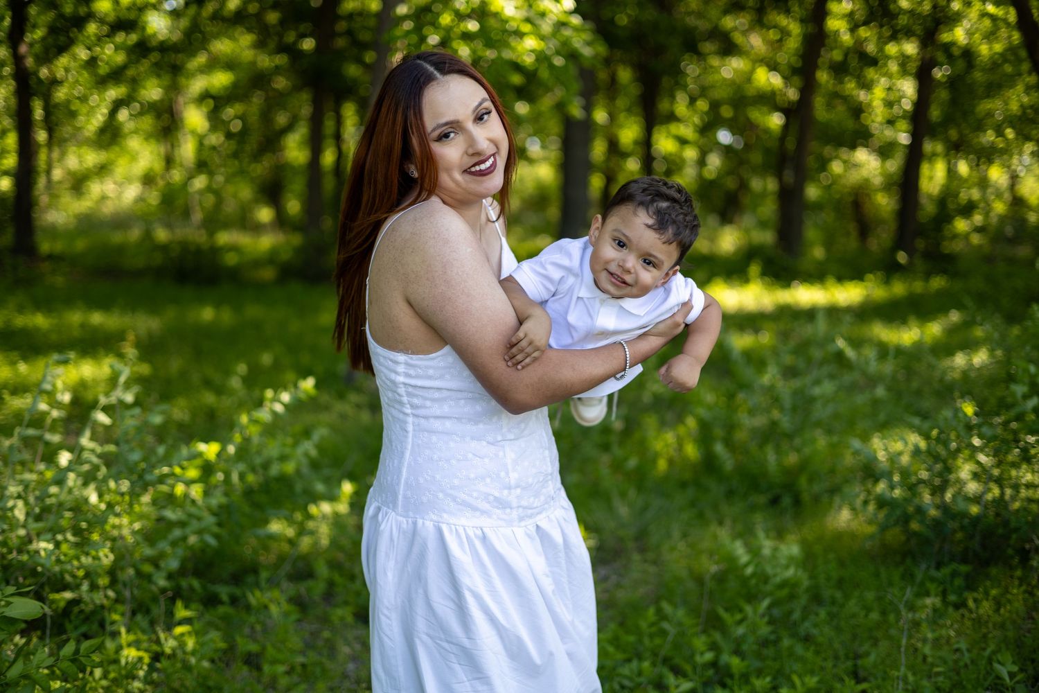 A smiling woman in a white dress holds a baby boy outdoors in a lush green park setting.