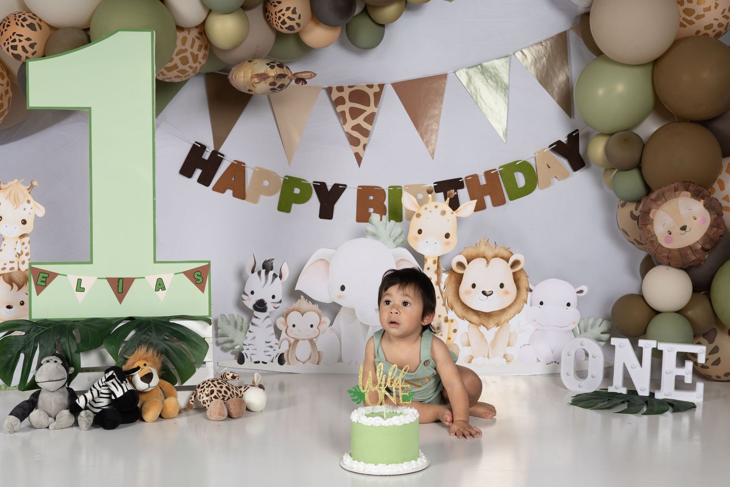 Baby boy sits before a green cake at a safari-themed first birthday party with animal toys and Happy Birthday banner.