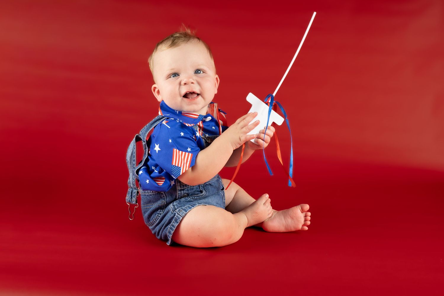 Smiling baby in patriotic American flag shirt and denim shorts sitting on red background holding a small white flag.