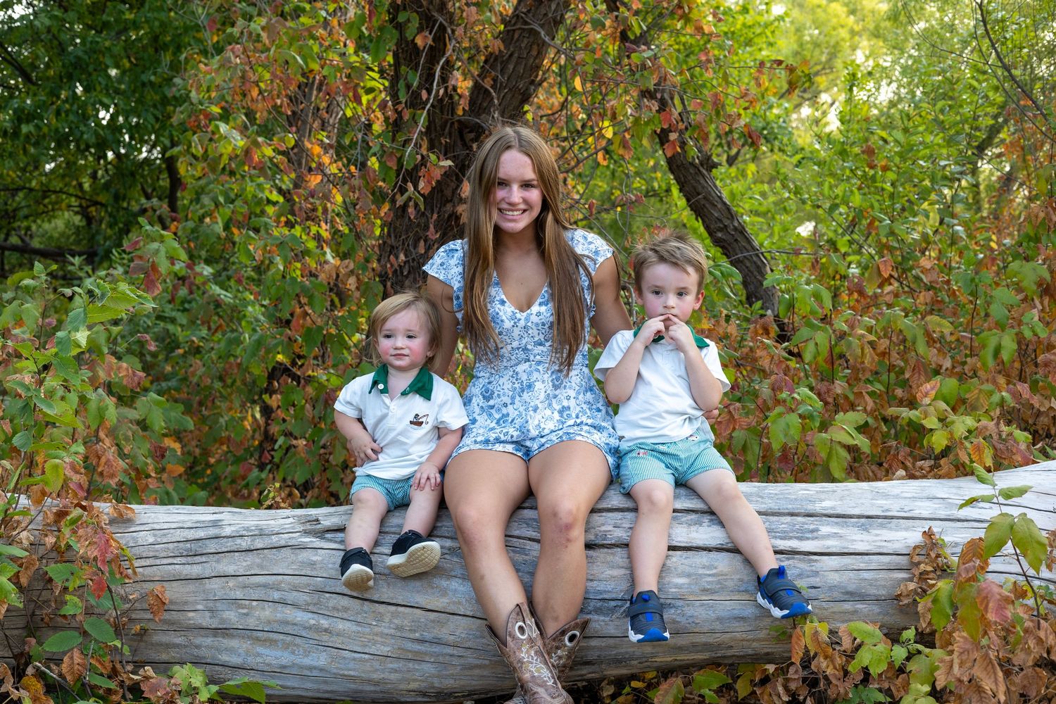 Woman and two toddlers sitting on a fallen log surrounded by lush green and autumn foliage outdoors.