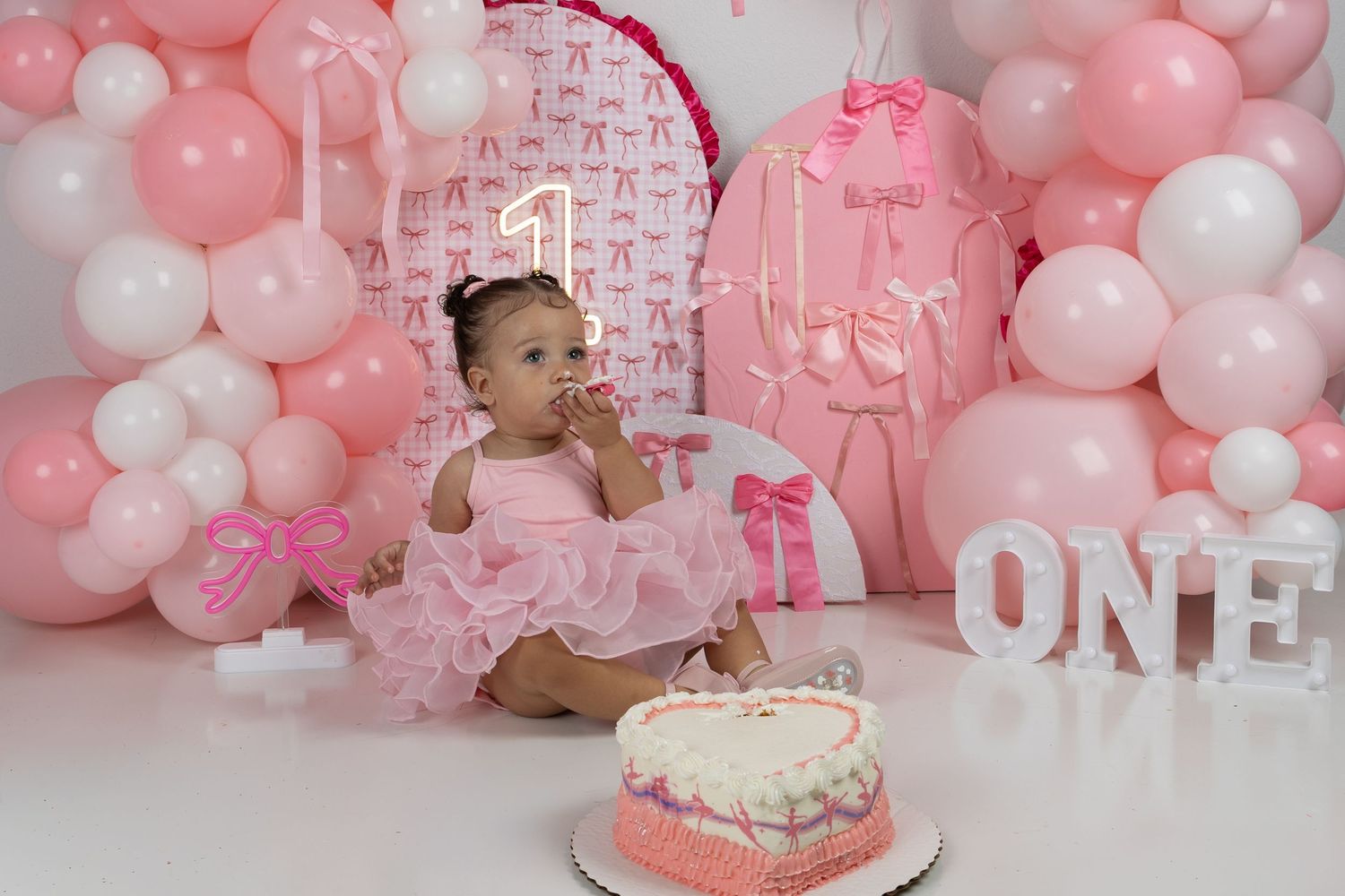 Baby girl in pink tutu sits before heart cake at a pink balloon first birthday smash cake photo session.