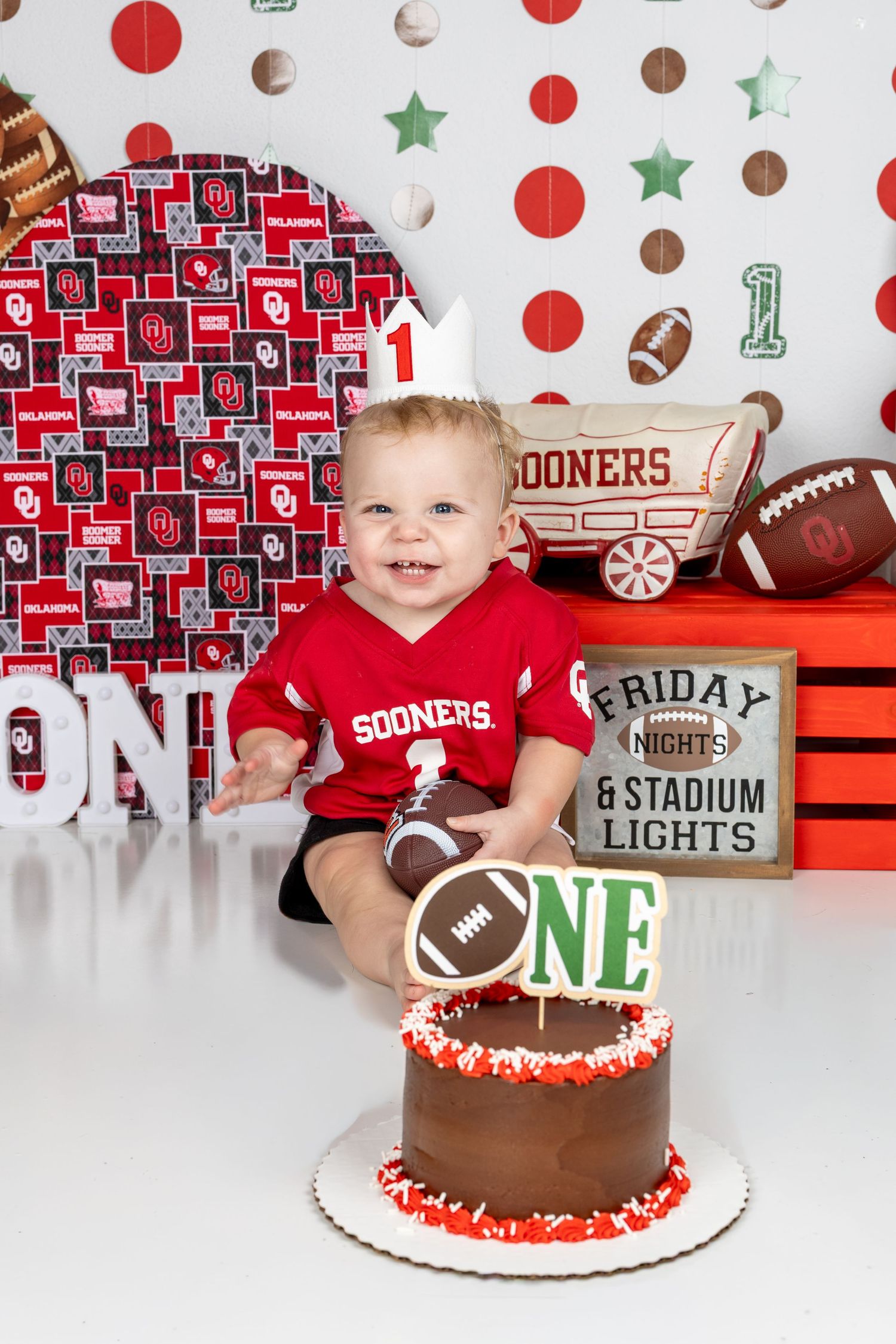 Baby in Sooners jersey celebrates first birthday with football-themed cake and Oklahoma Sooners decorations.