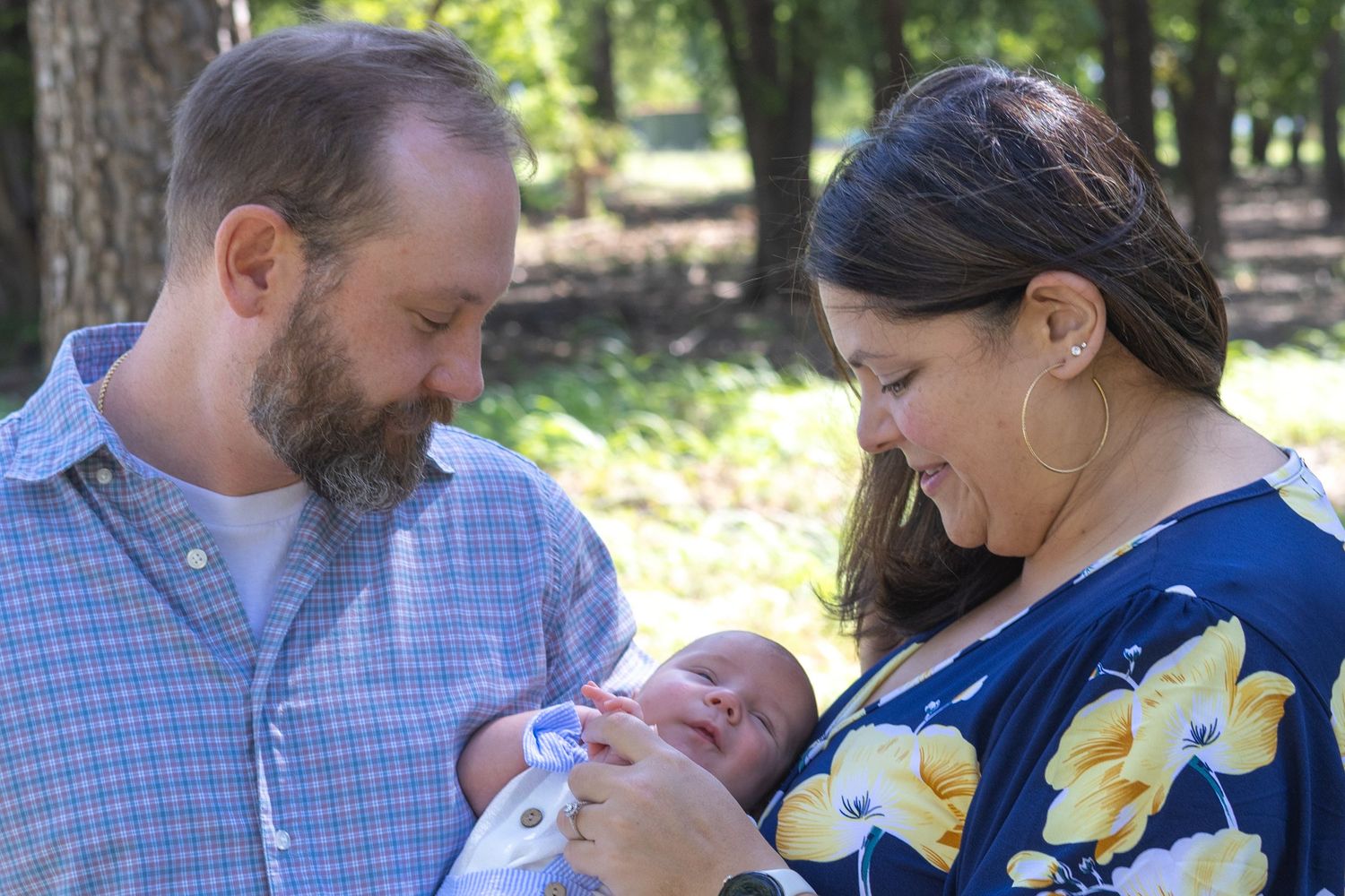 A couple admiring their newborn baby outdoors among trees, woman in floral top, man in blue shirt.