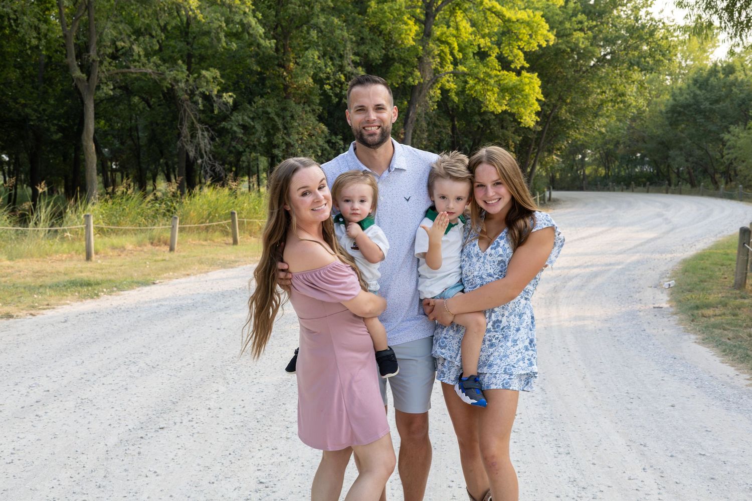 Family of five posing together on a gravel road surrounded by lush green trees on a sunny day.