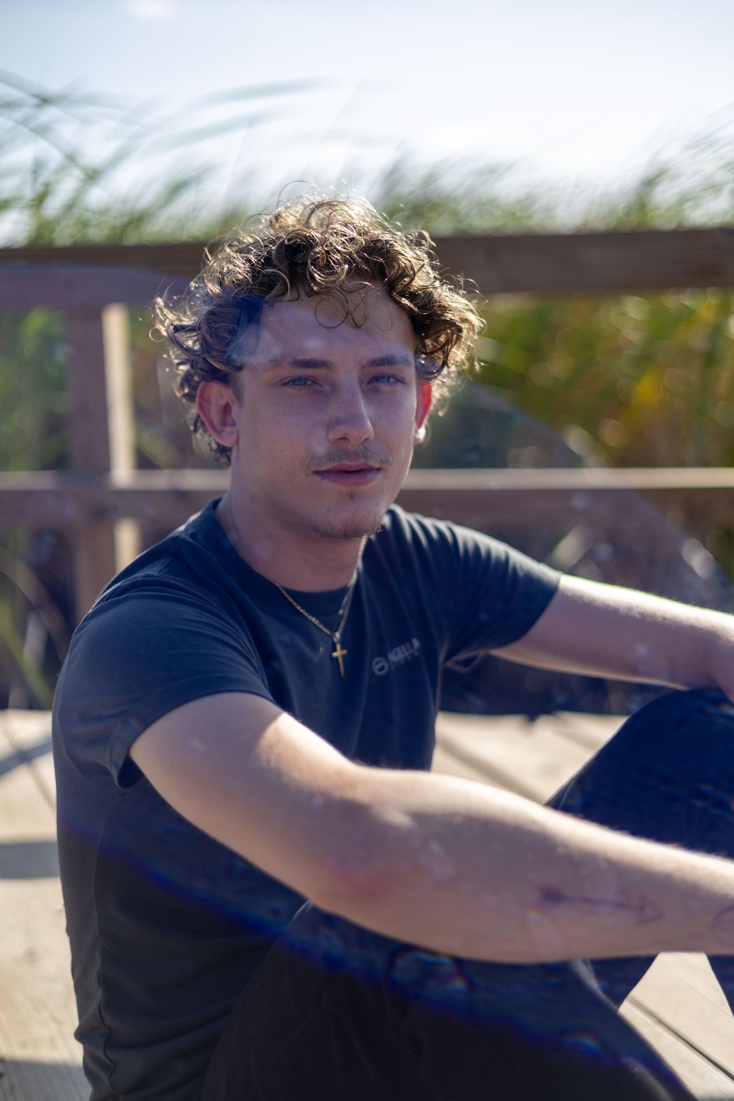 Young man with curly hair wearing a black t-shirt and gold cross necklace sitting on a wooden deck outdoors.
