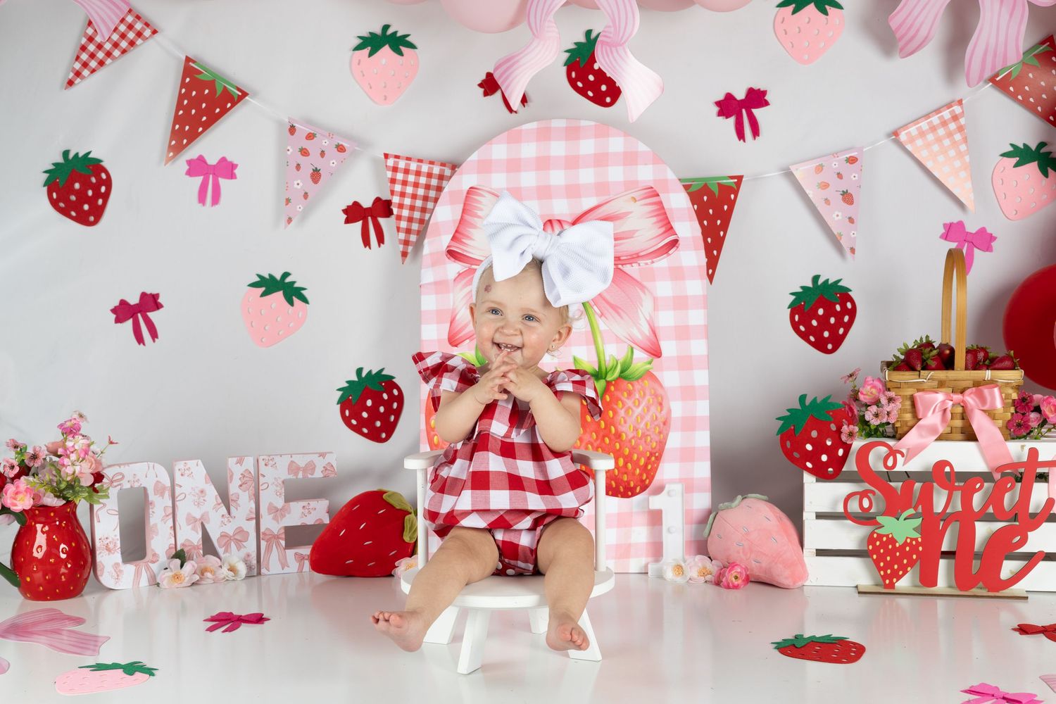 Baby girl in red gingham outfit sits in strawberry-themed first birthday cake smash setup with ONE letters and decorations.
