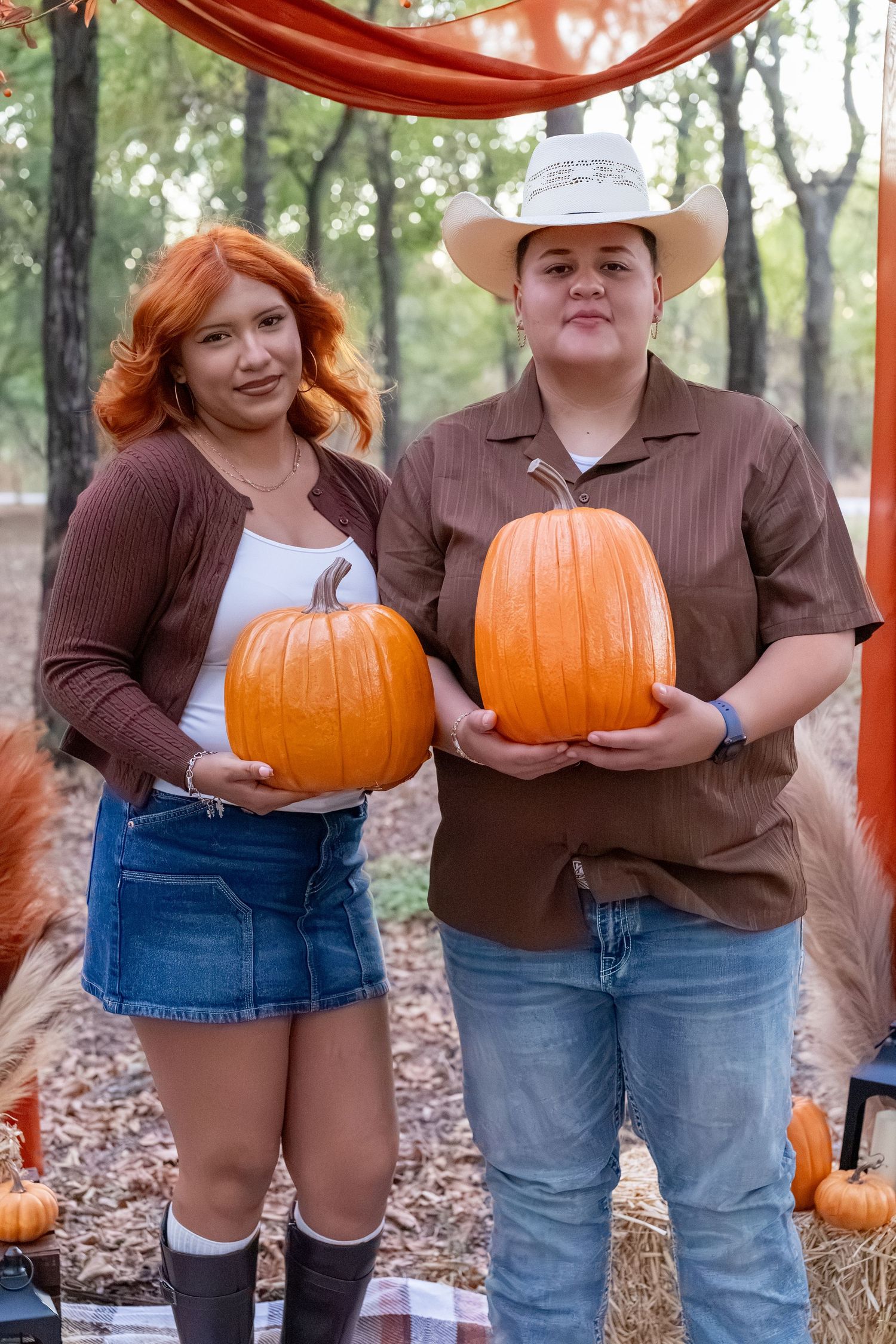 Two women holding large orange pumpkins outdoors at a fall harvest festival, surrounded by hay bales and autumn decorations.