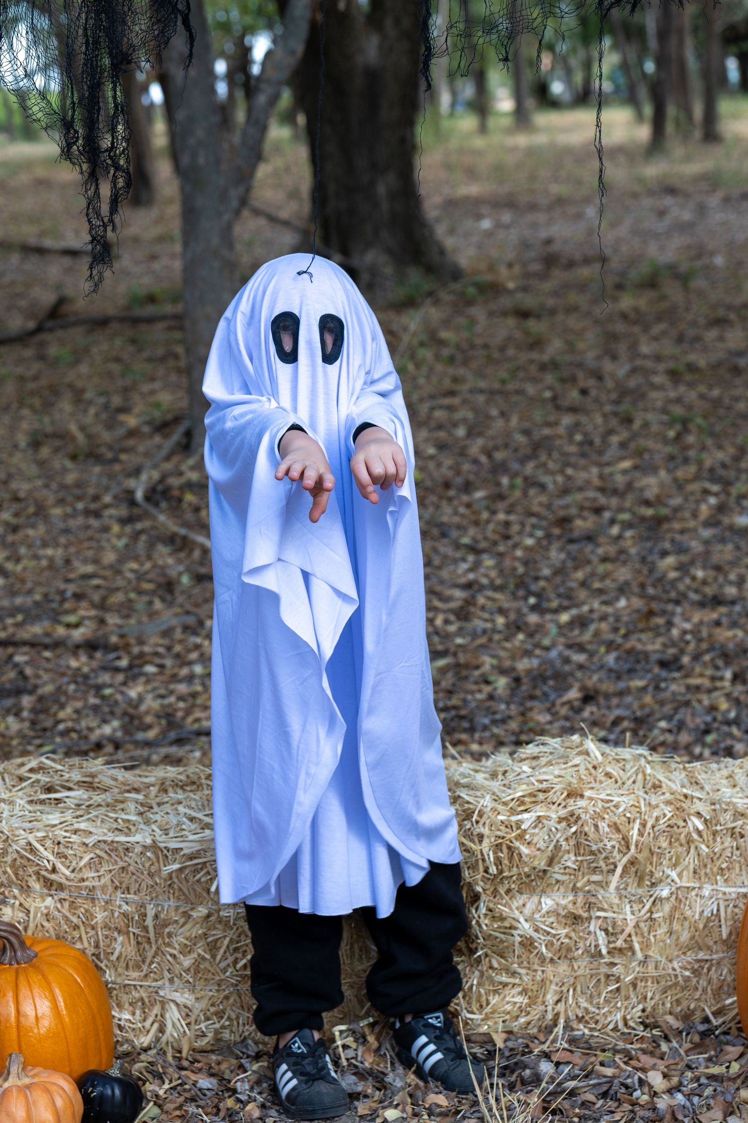 Child in white ghost Halloween costume with black eyes stands by hay bales and pumpkins in wooded outdoor setting.