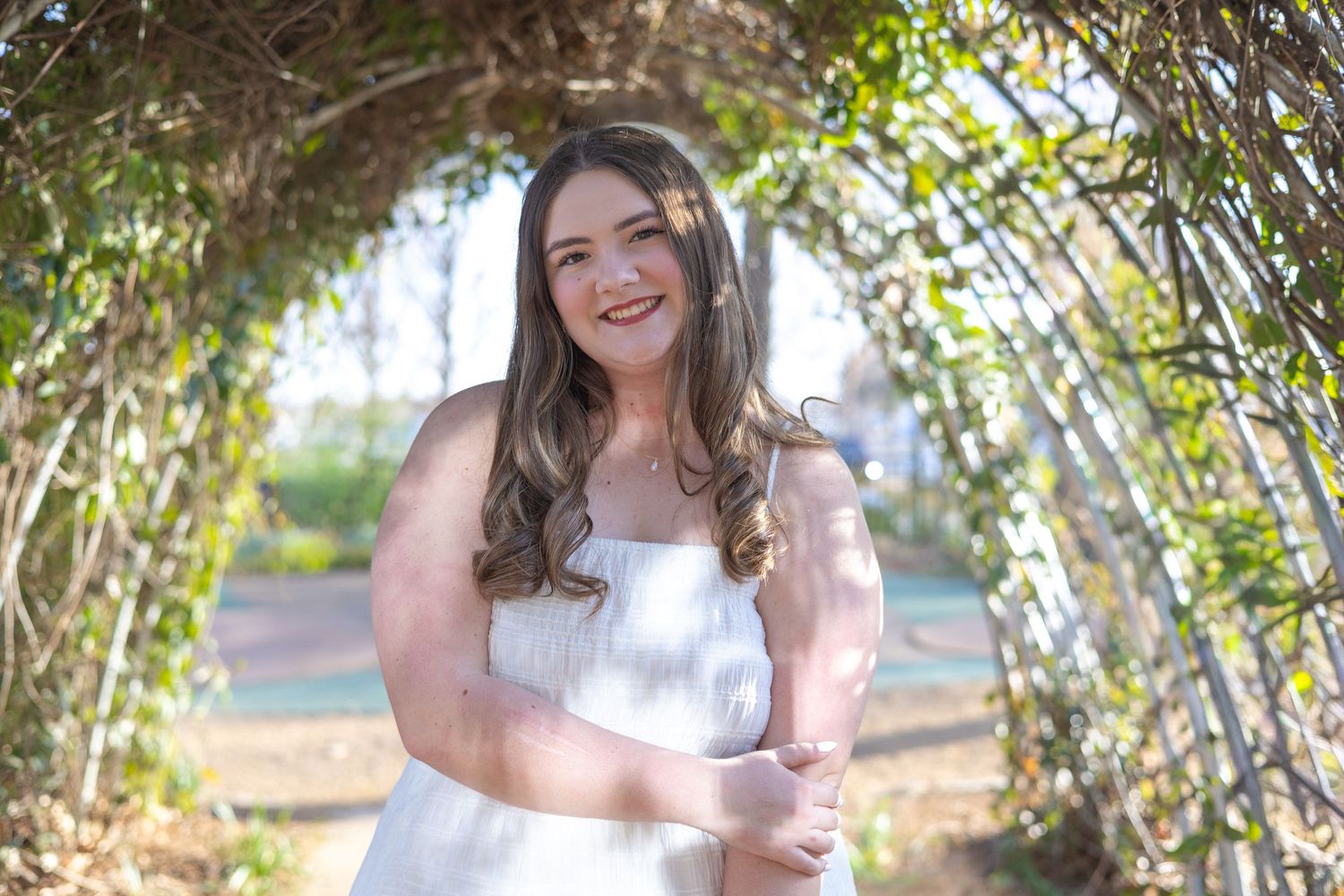 A person in a white dress poses outdoors among sunlit trees and greenery during a photoshoot session.