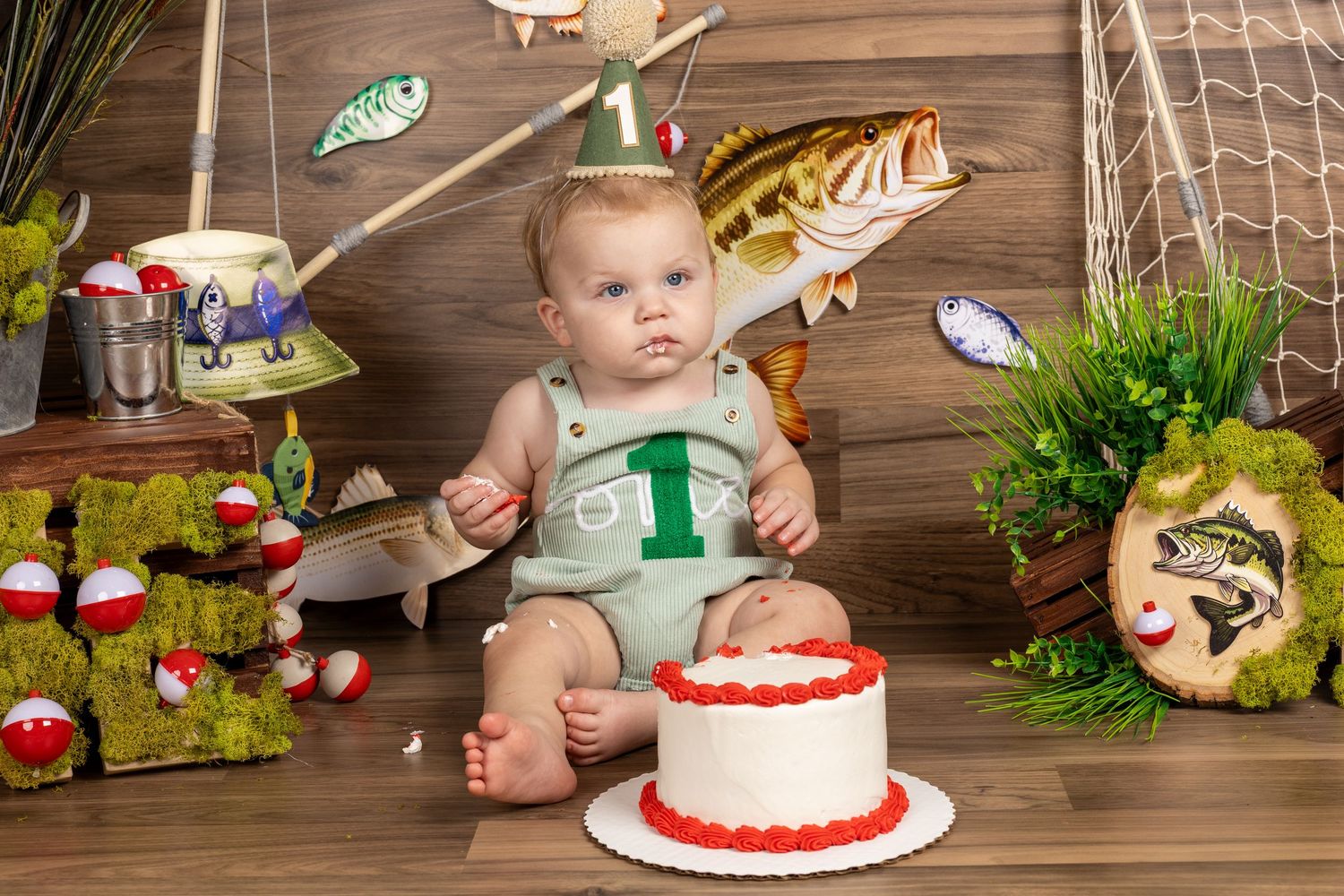Baby in green fishing-themed outfit sits beside a white cake celebrating his 1st birthday with fish decor.