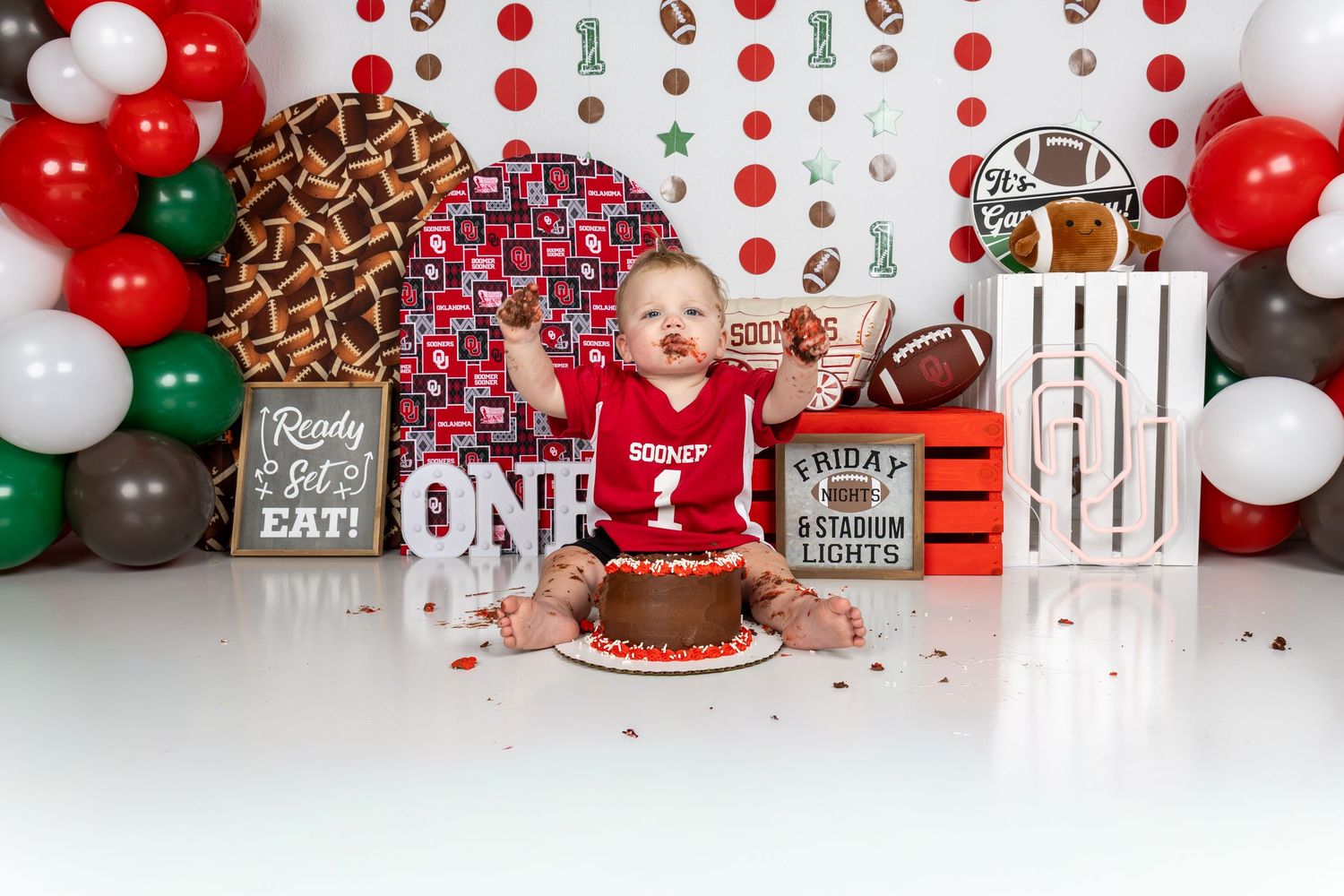 Baby boy in red jersey smashes first birthday cake at football-themed cake smash with balloons and sports decor.