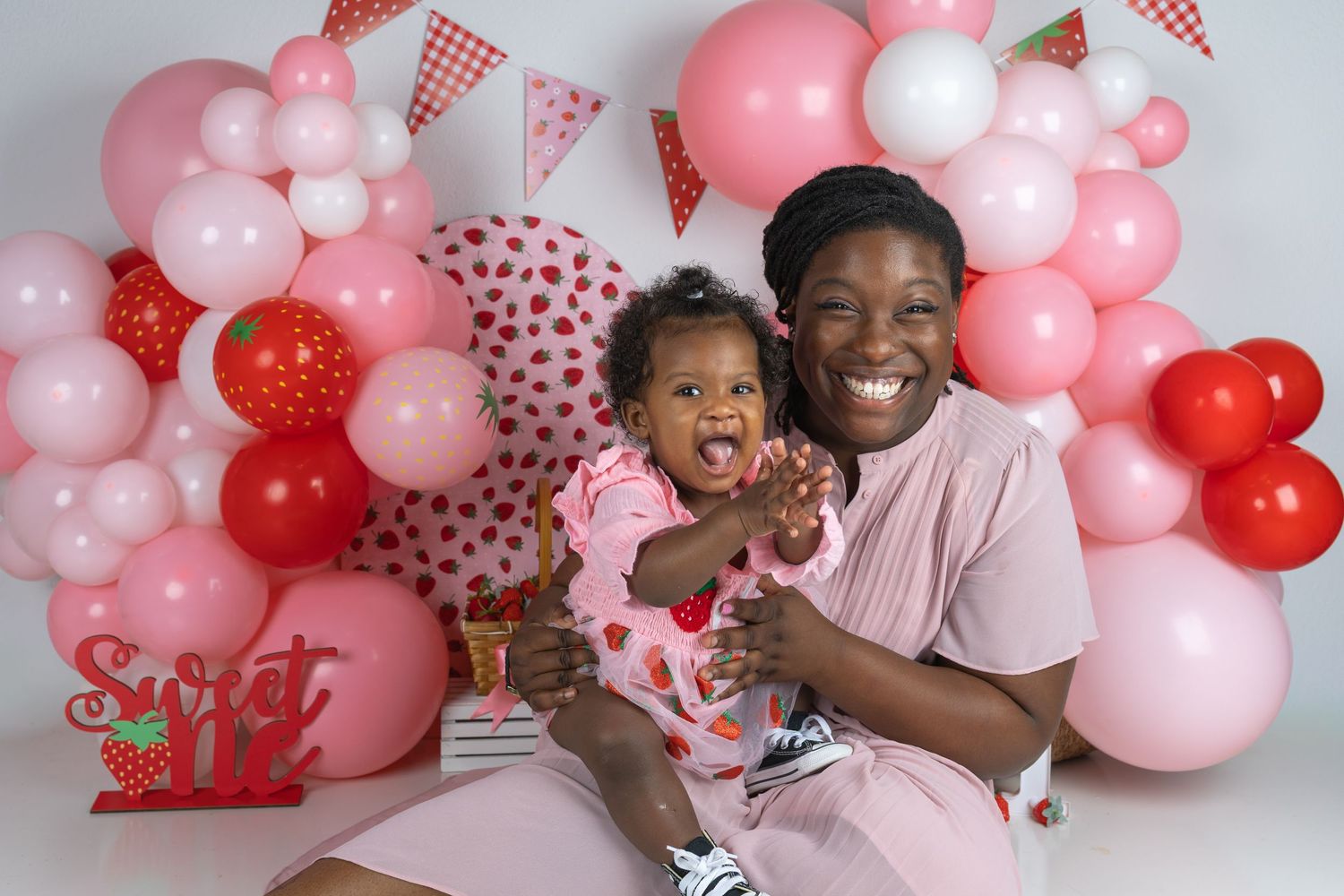A mother holds her baby at a colorful pink and red balloon backdrop birthday photo shoot.