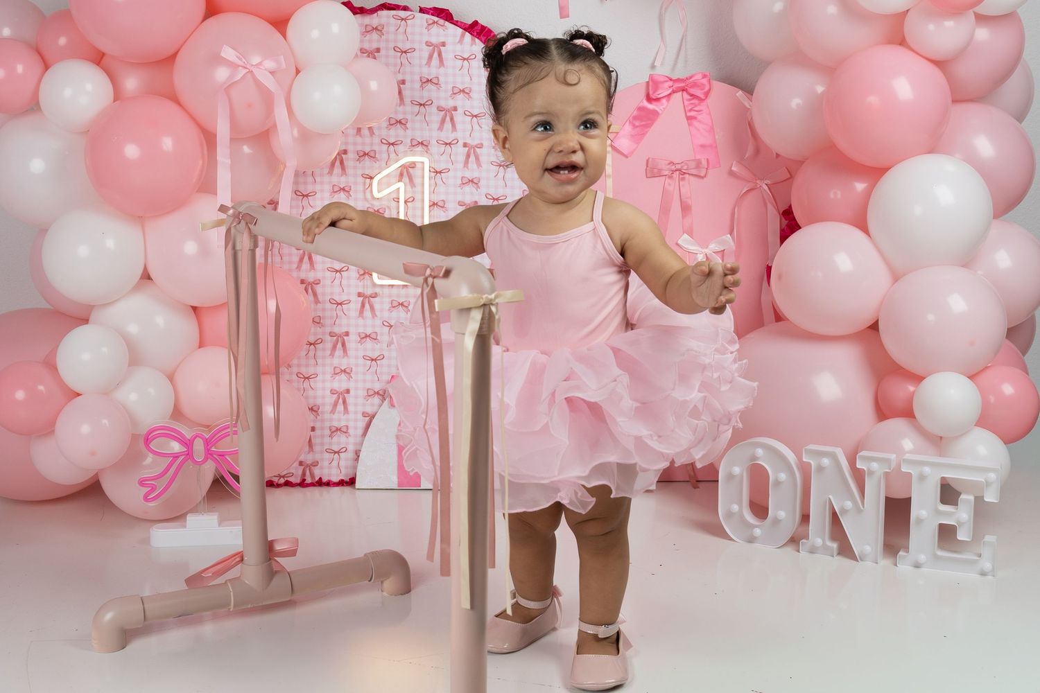A baby girl in a pink tutu dress stands at a ballet barre at her first birthday party with pink balloon decorations.