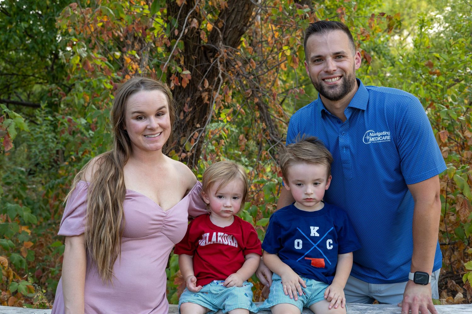 A smiling family of four poses outdoors in autumn, with two toddlers seated between their parents near colorful foliage.