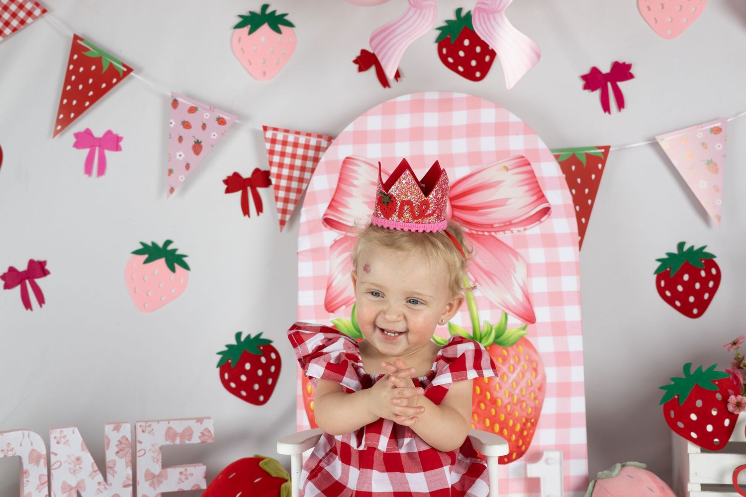 Smiling toddler girl in strawberry dress with crown, surrounded by strawberry-themed birthday decorations and bunting.