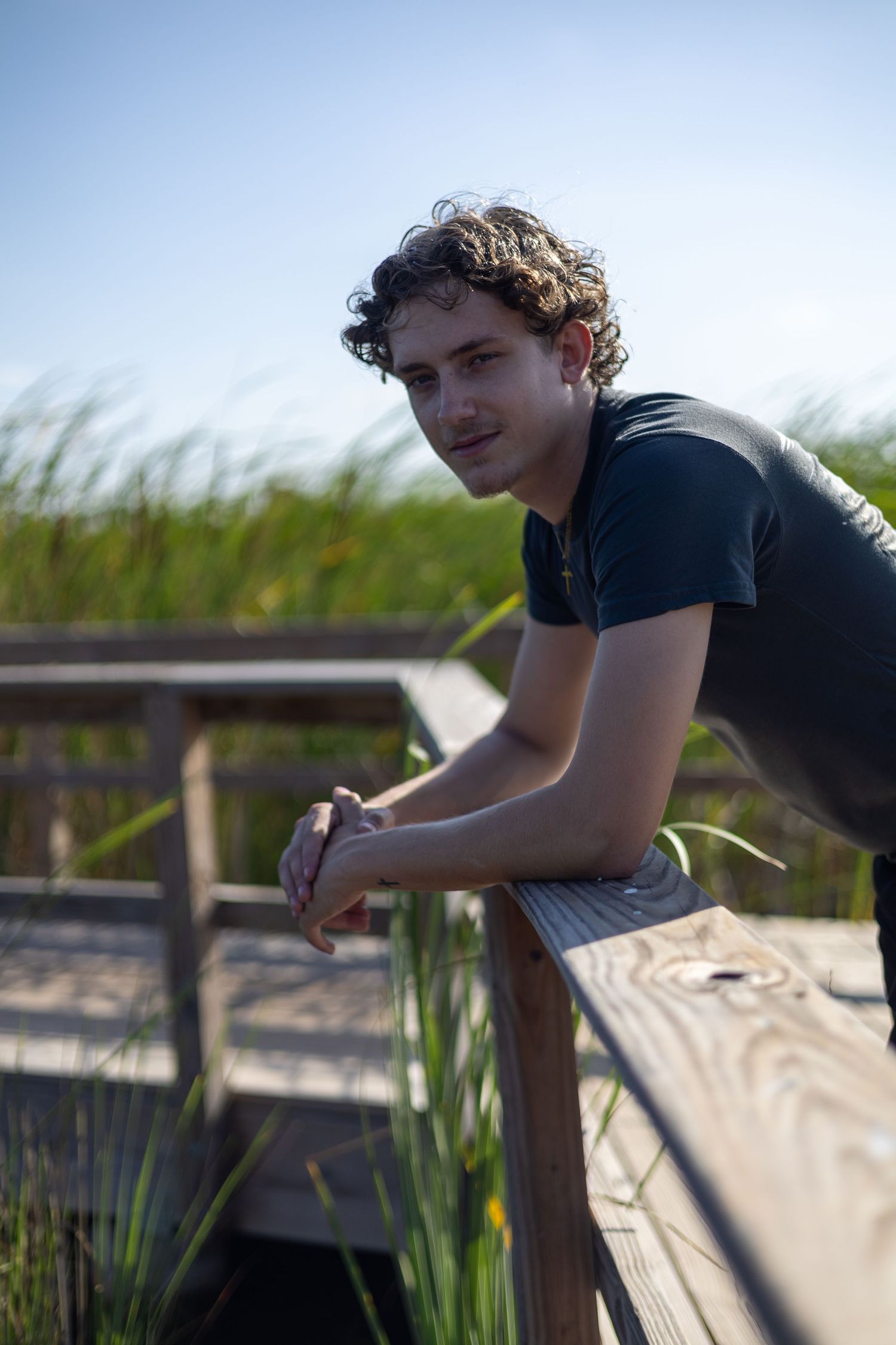 Young man with curly hair leans on wooden boardwalk railing outdoors, green marsh grass and blue sky behind him.