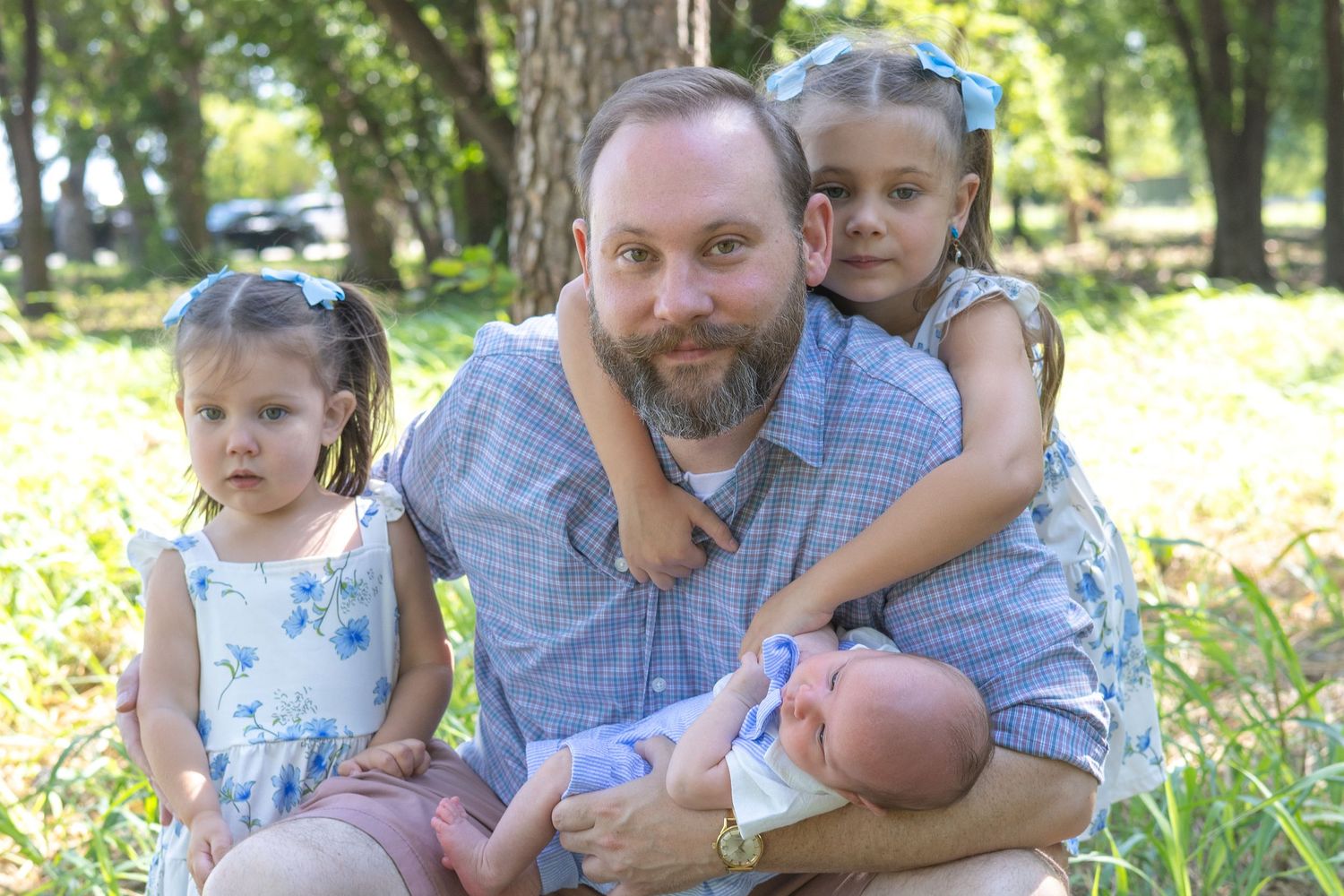A bearded father sits outdoors holding a newborn baby with two young girls in matching floral dresses beside him.