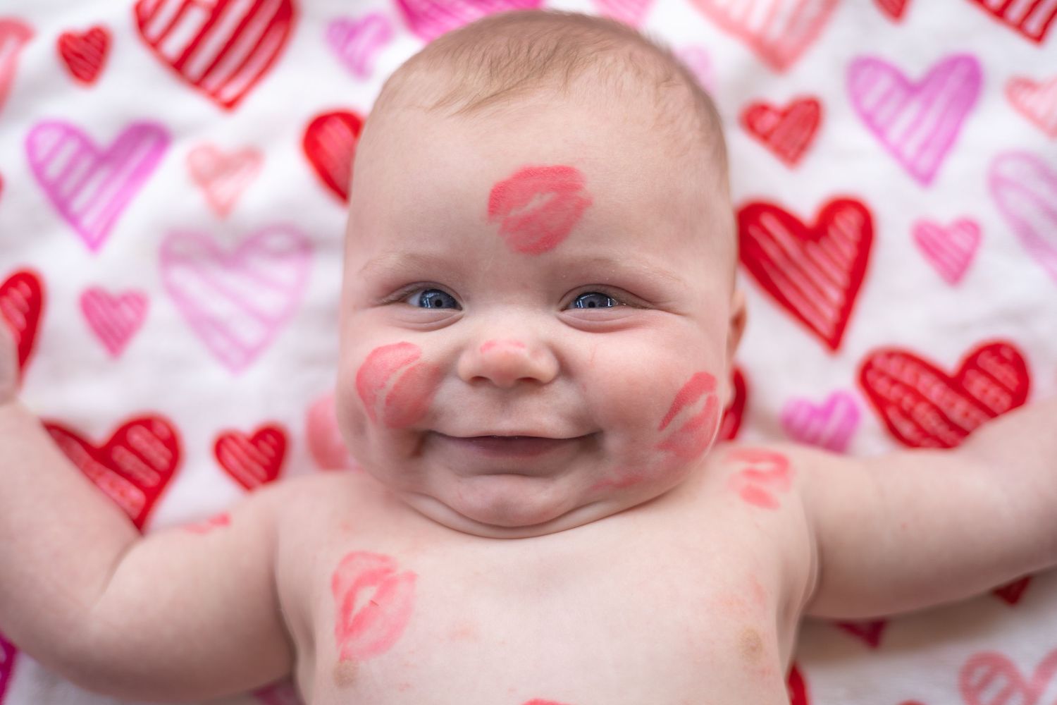 Baby lying on white surface with pink and red heart pattern background smiling joyfully.