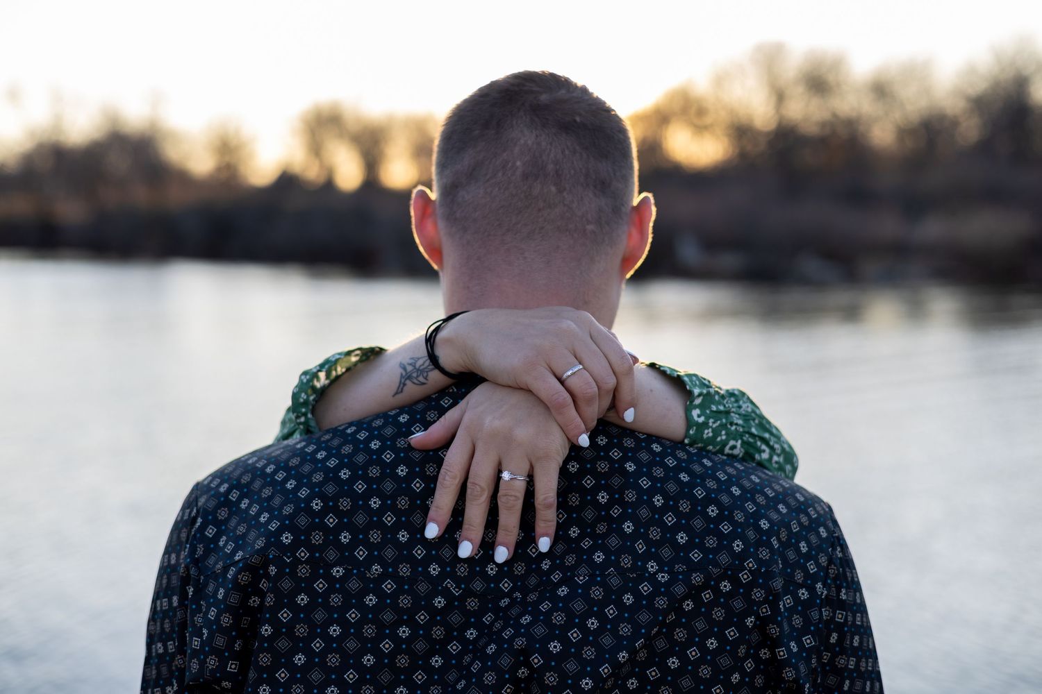 Someone wearing a polka dot sweater being embraced from behind with hands visible at sunset by a lake.