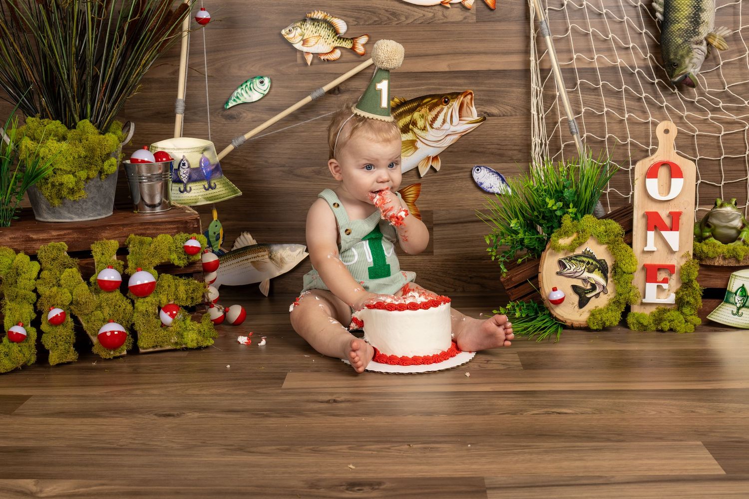 Baby enjoys first birthday cake smash with fishing theme decor, moss letters, and fish decorations on wood backdrop.