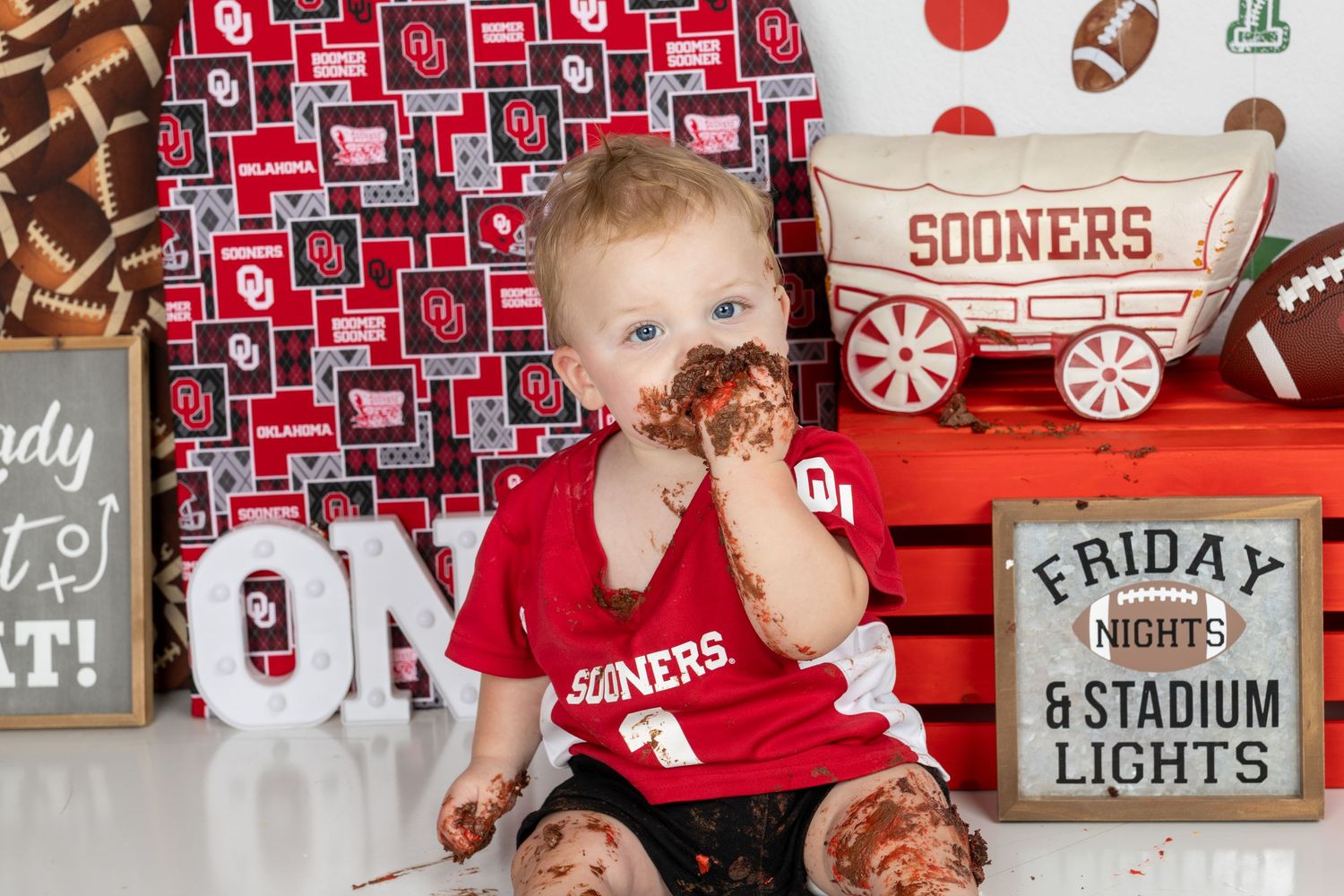 Baby in Sooners jersey enjoys smash cake at Oklahoma football-themed first birthday party with OU decorations.
