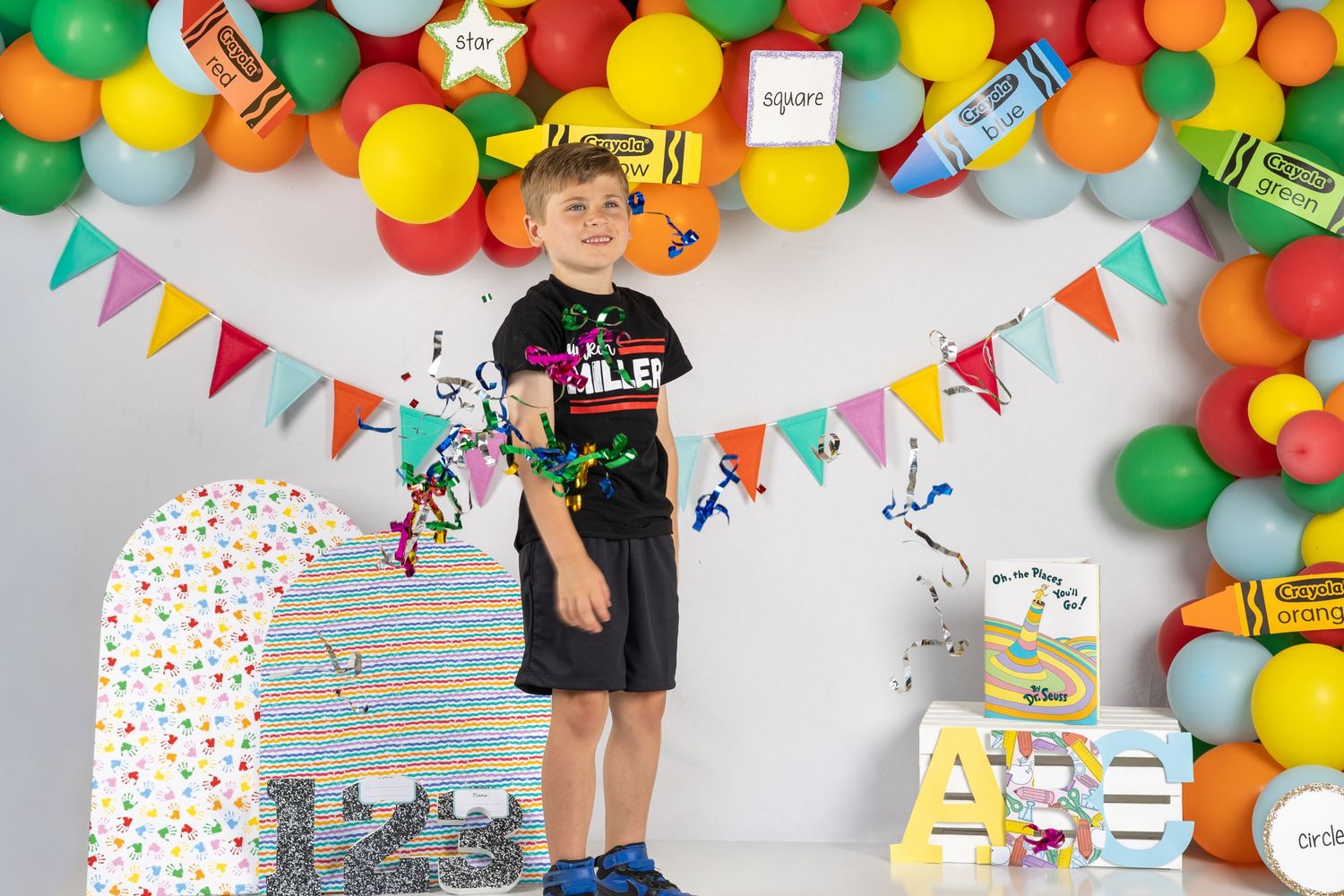 Boy stands at colorful school-themed birthday party backdrop with balloons, pennants, and educational decorations.