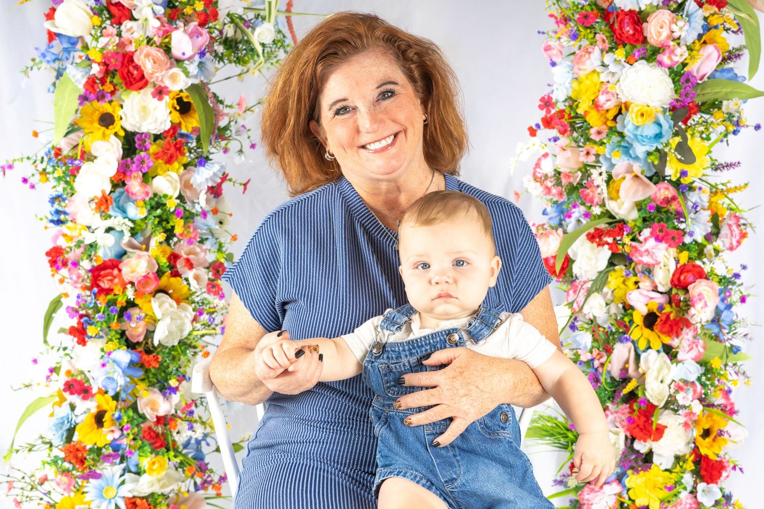 Woman and baby sitting on a floral swing surrounded by colorful flowers, smiling together in a bright studio setting.