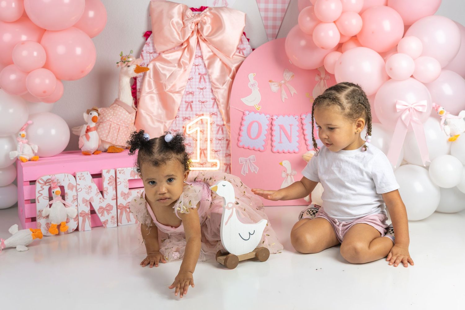 Two baby girls celebrate a first birthday with pink balloons, ONE letters, and a rocking horse in a pink party setup.