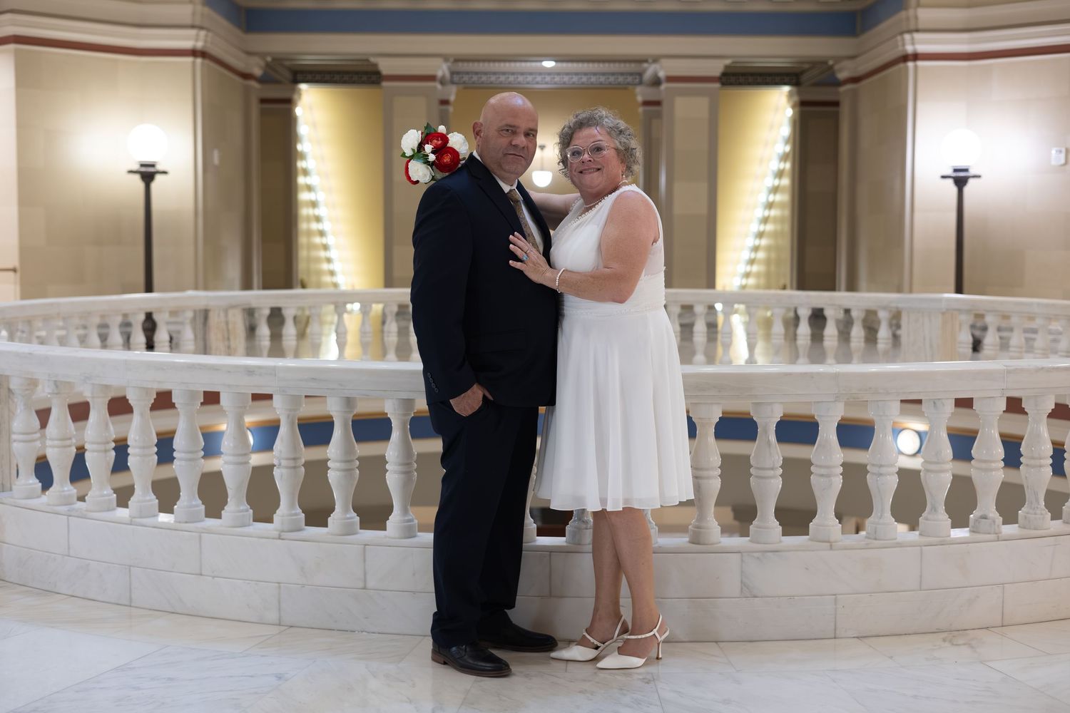 Couple in wedding attire posing by white marble staircase railing in elegant indoor venue.