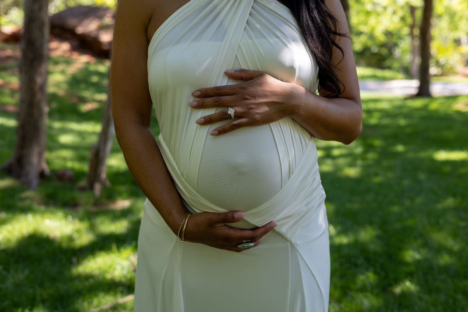 Pregnant woman in white halter dress cradling baby bump outdoors in a green park setting.