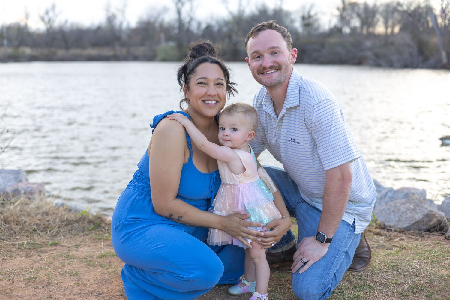 Family in coordinating blue and white outfits poses together by a lake during sunset for outdoor portraits.