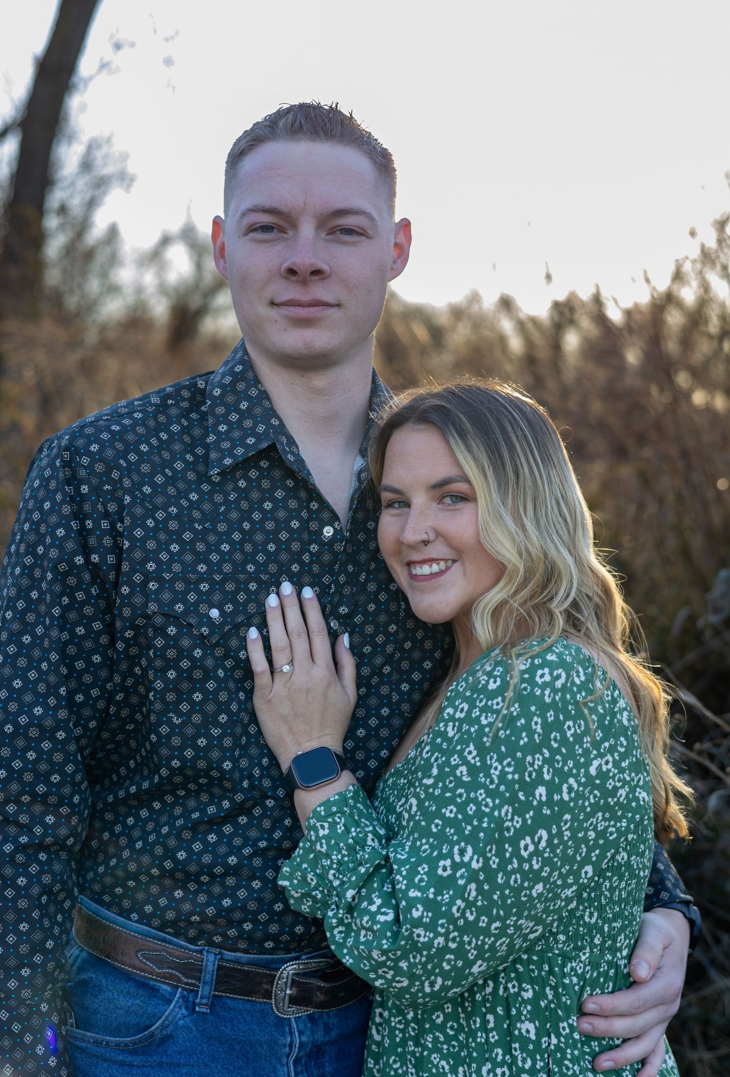 A couple poses together outdoors during an engagement photo session, with the person in green showing off their ring.