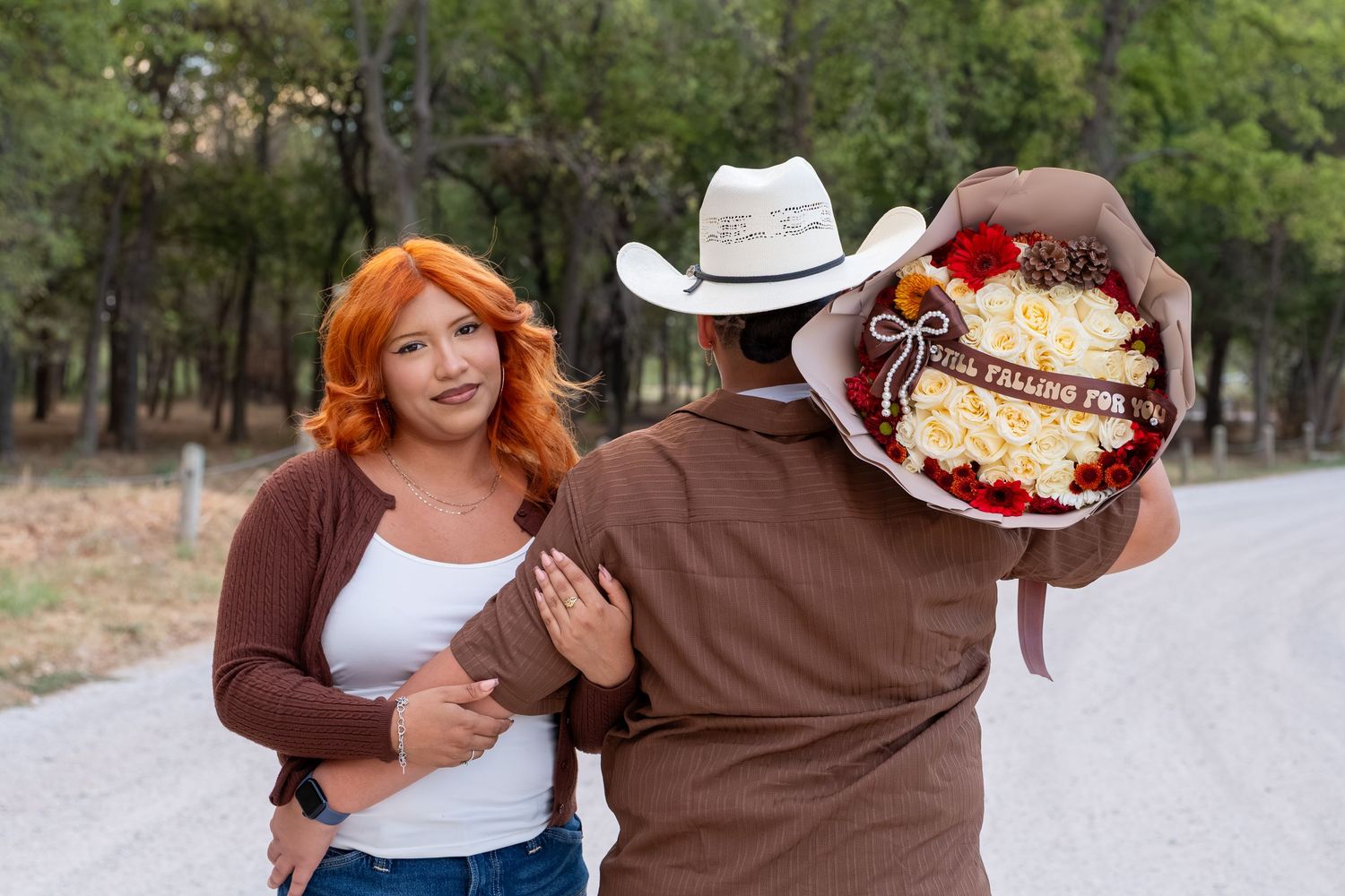 Man in cowboy hat surprising red-haired woman with large floral bouquet on a scenic country road.