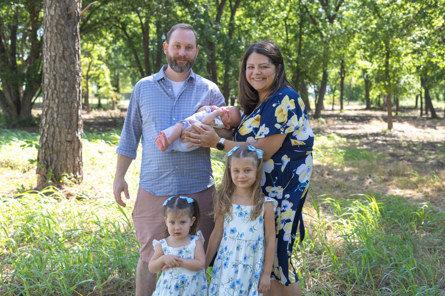 Family of five posing outdoors in a lush green park, parents holding newborn baby with two young daughters in matching dresses.
