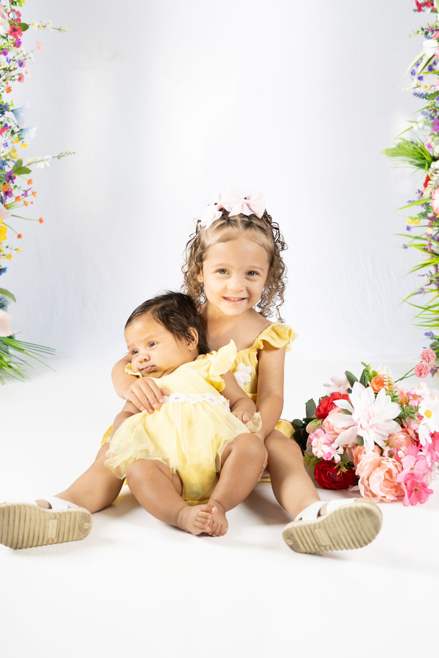Two young sisters in yellow dresses pose together surrounded by colorful flowers on a white background.