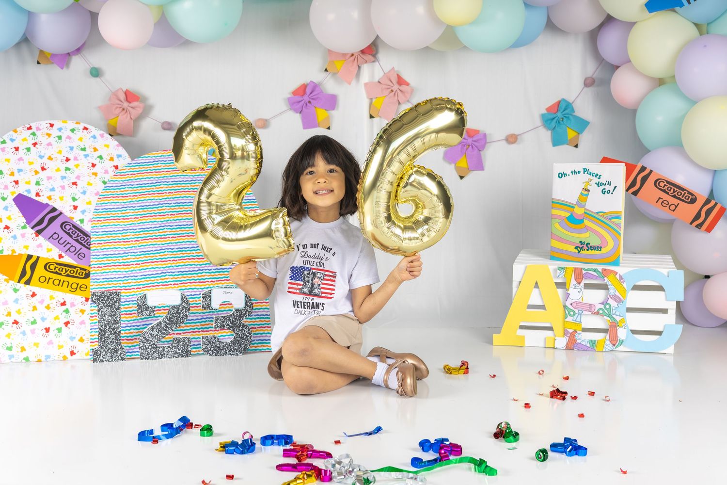 A young girl holds gold number 26 balloons at a colorful birthday party setup with pastel balloons and school-themed decorations.