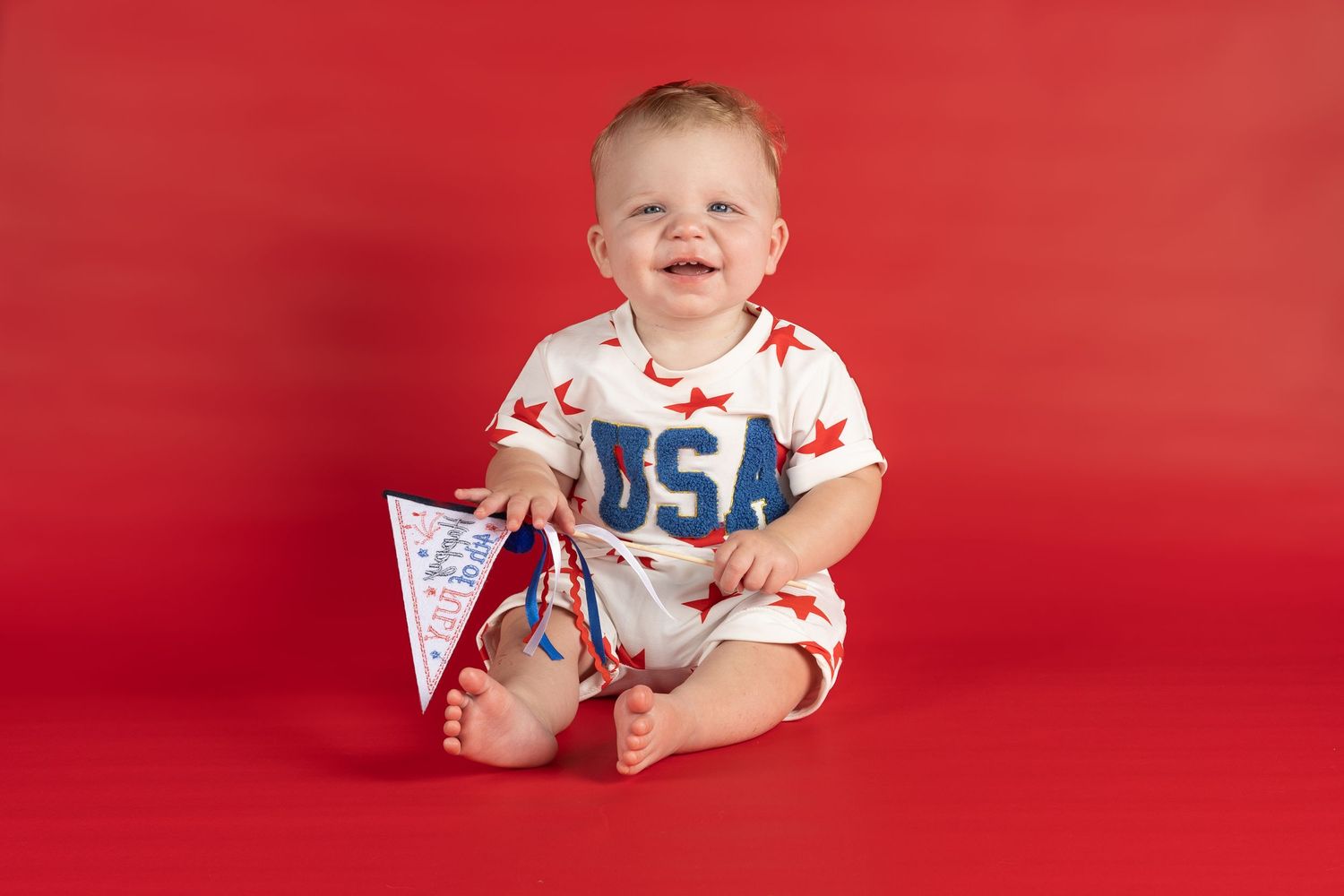 Smiling baby in a USA romper with red stars holds a pennant flag against a bright red background.