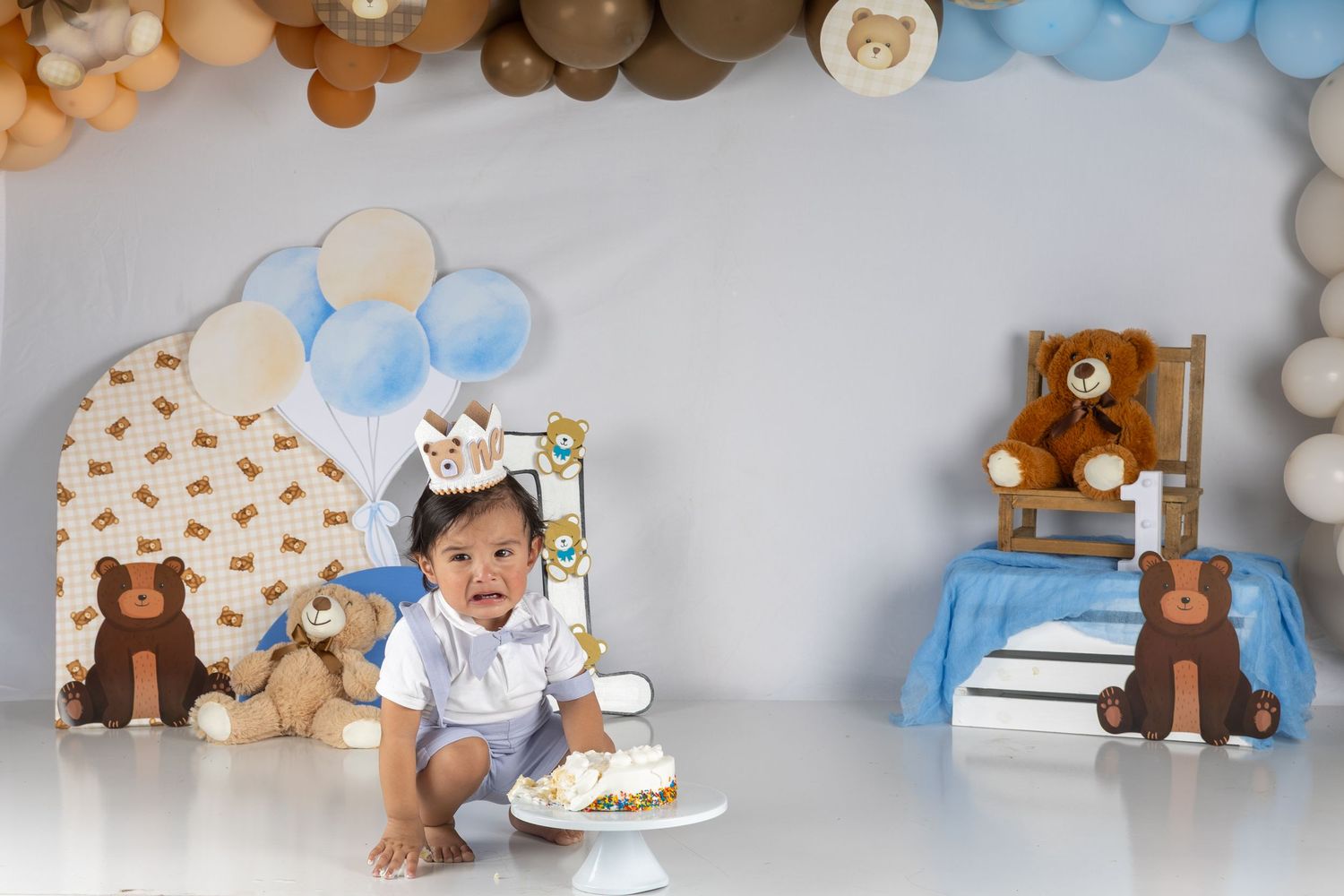Baby in crown sits with smash cake at teddy bear first birthday party setup with balloons and decorations.