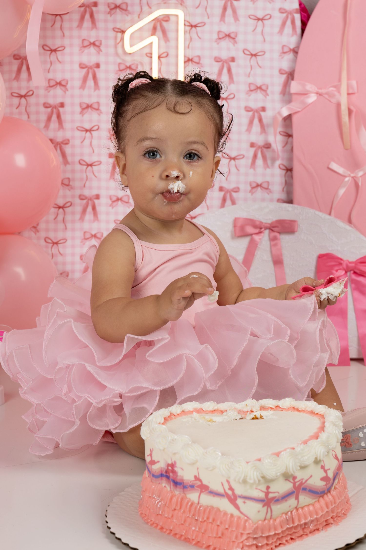 Baby girl in pink tutu celebrating first birthday with heart-shaped cake, frosting on face, surrounded by pink balloons and bows.