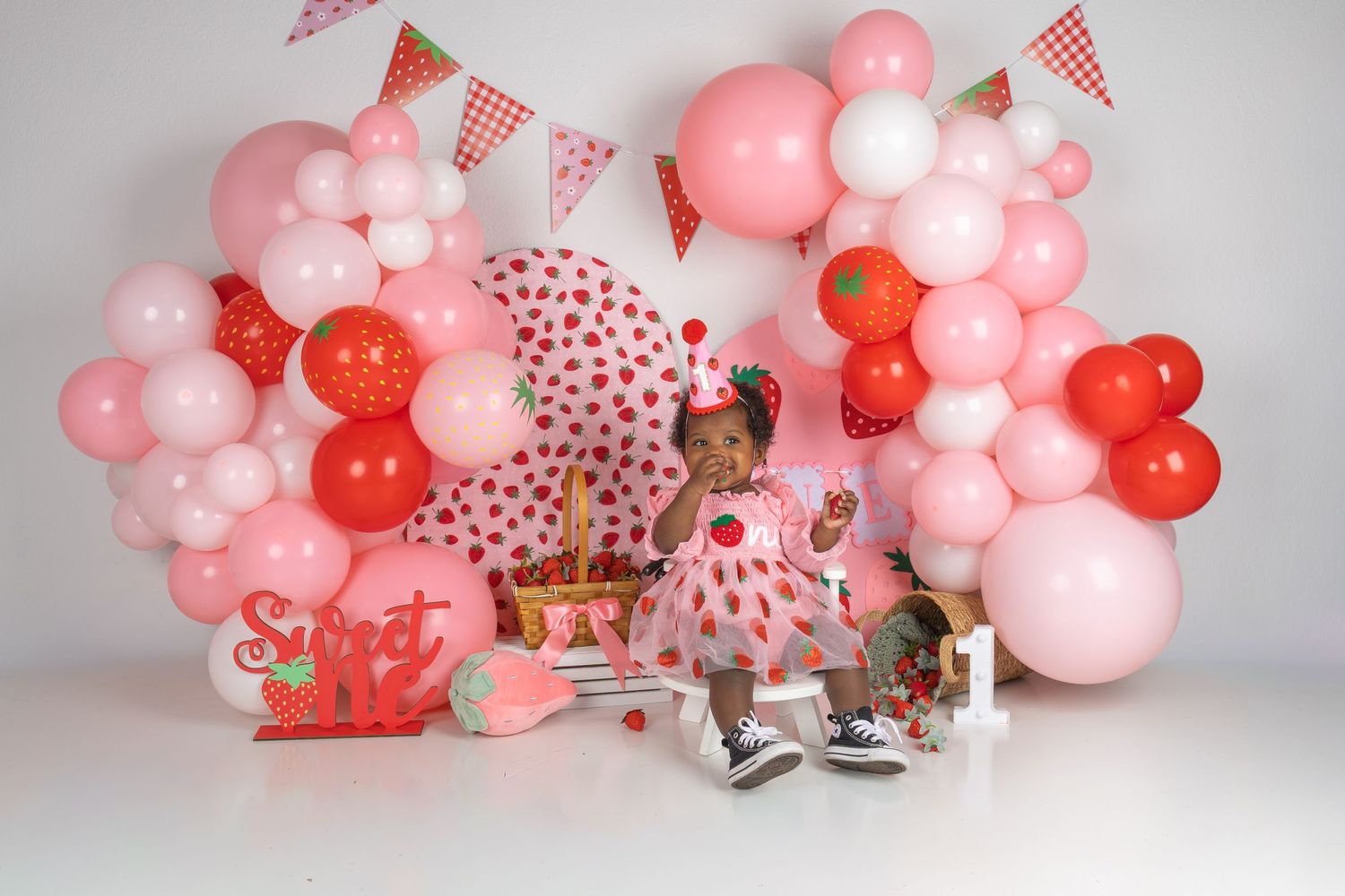 Baby girl in strawberry dress celebrates first birthday with pink and red balloon arch and sweet one decorations.