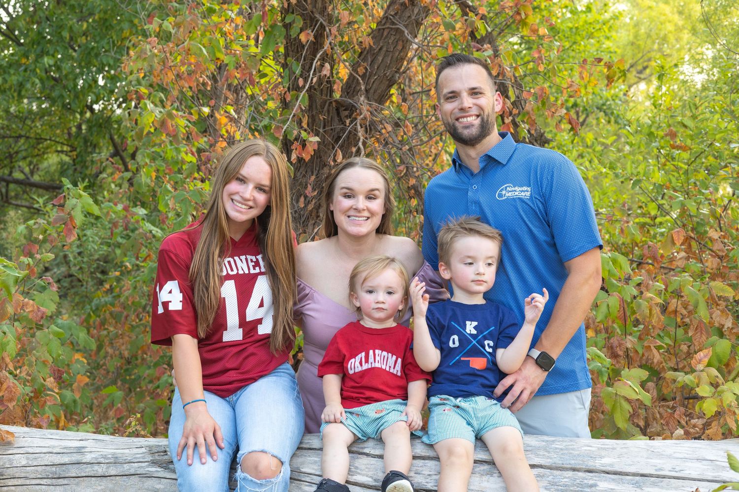 Family of five smiling outdoors on a log, wearing Oklahoma Sooners jerseys, surrounded by colorful autumn foliage.