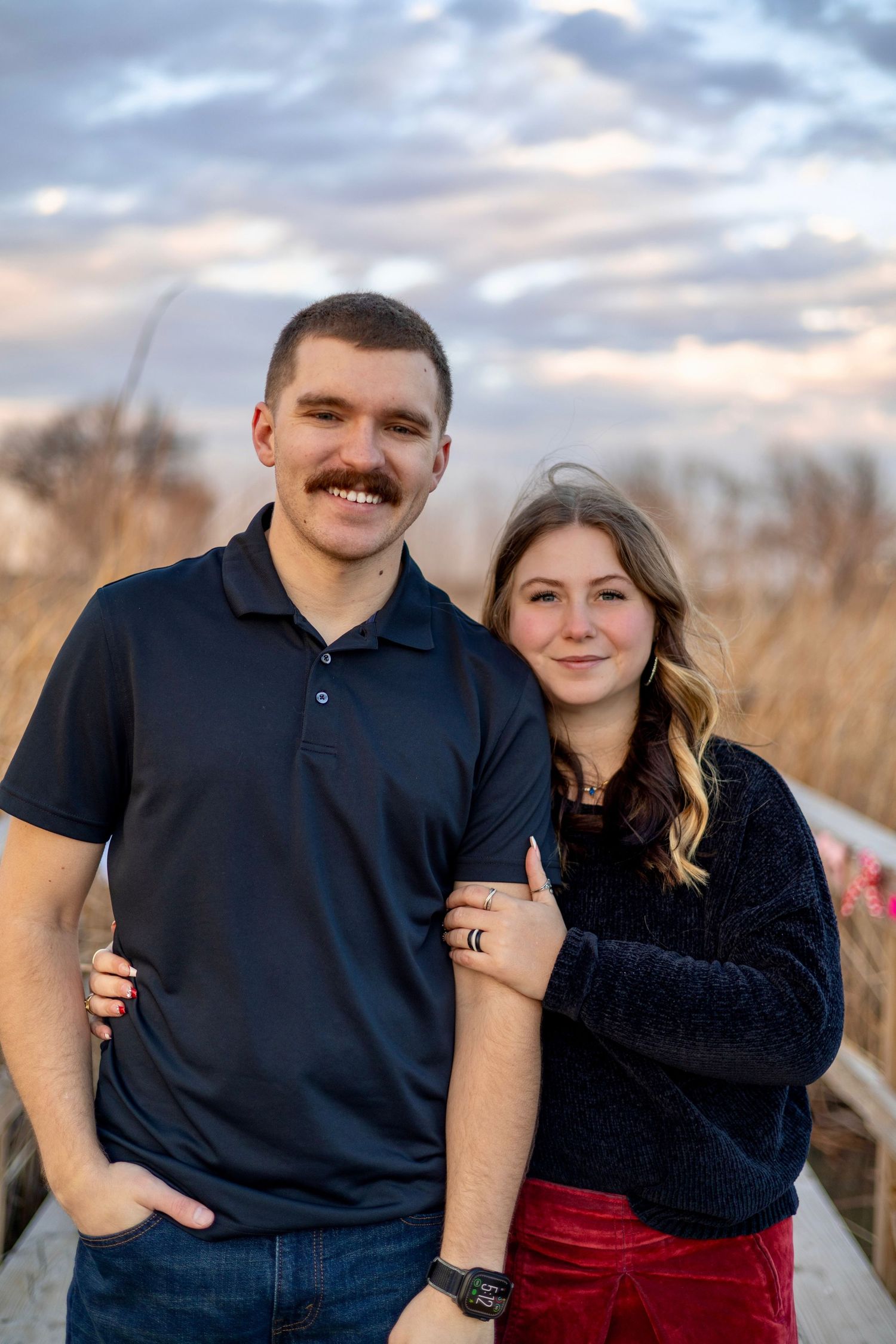 A couple poses together in a field at sunset wearing matching dark tops and casual attire.