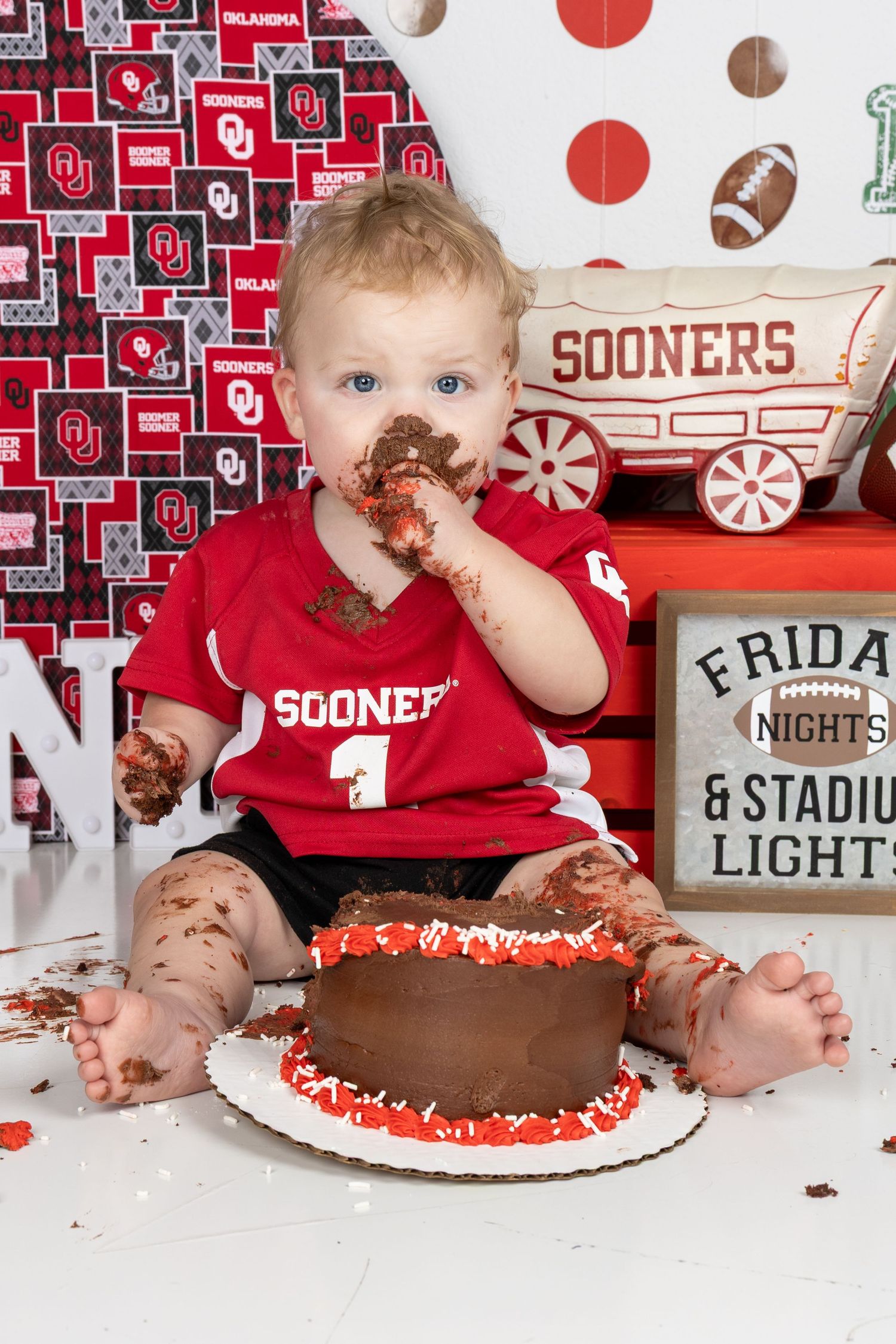 Baby in Sooners jersey smashes chocolate birthday cake during Oklahoma Sooners themed first birthday cake smash photo session.