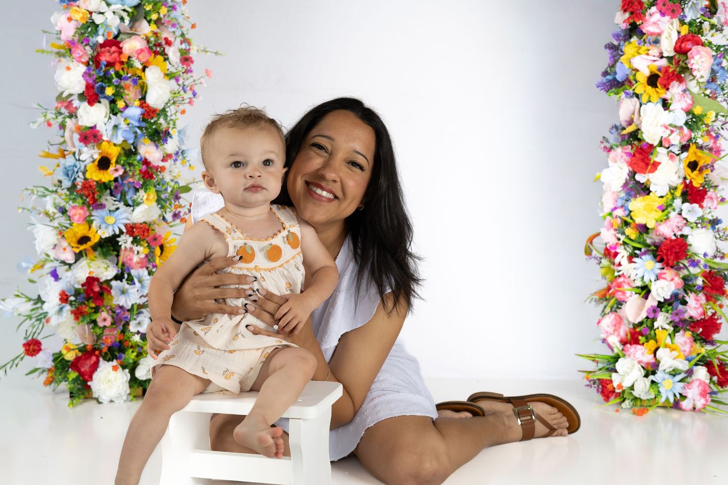 A smiling woman sits on a white floor holding a baby girl, framed by colorful floral columns on a white background.