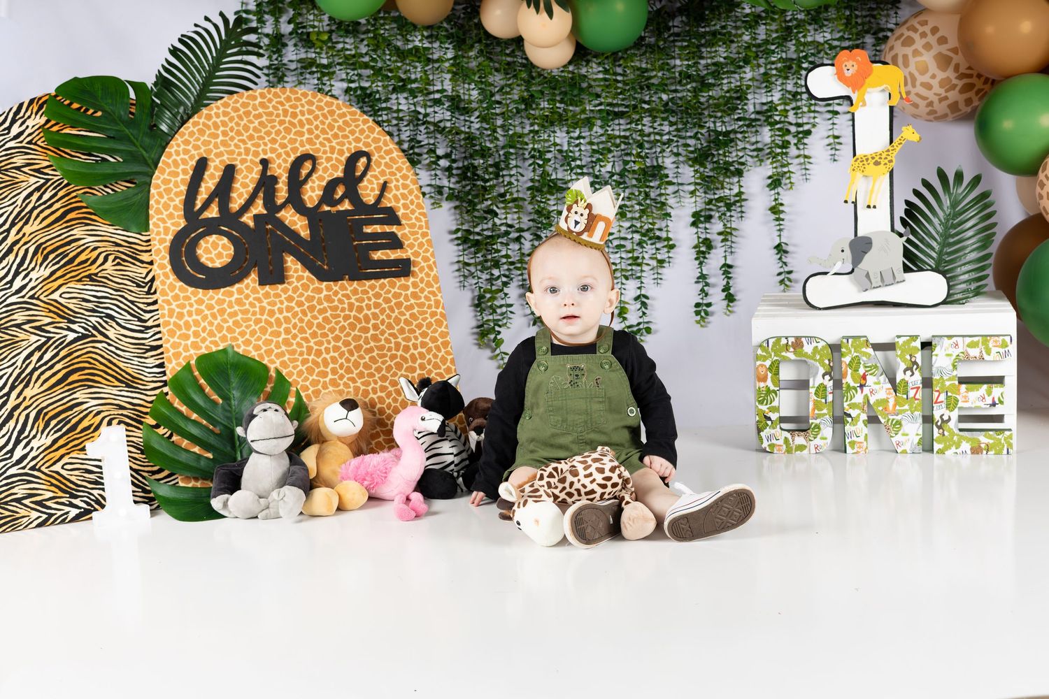 Baby in jungle-themed first birthday setup with stuffed animals, Wild One sign, and colorful balloon backdrop.